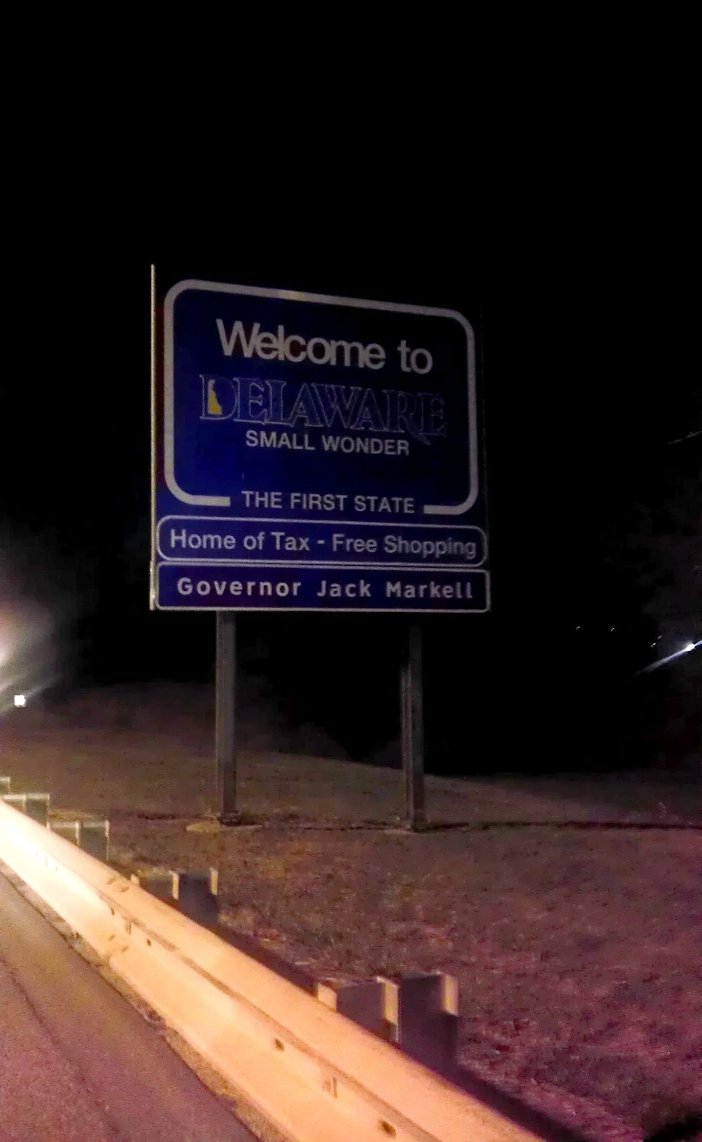 Nighttime photo of a welcome sign reading "Welcome to Delaware, Small Wonder, The First State, Home of Tax-Free Shopping, Governor Jack Markell" along a roadside with a safety barrier in the foreground.