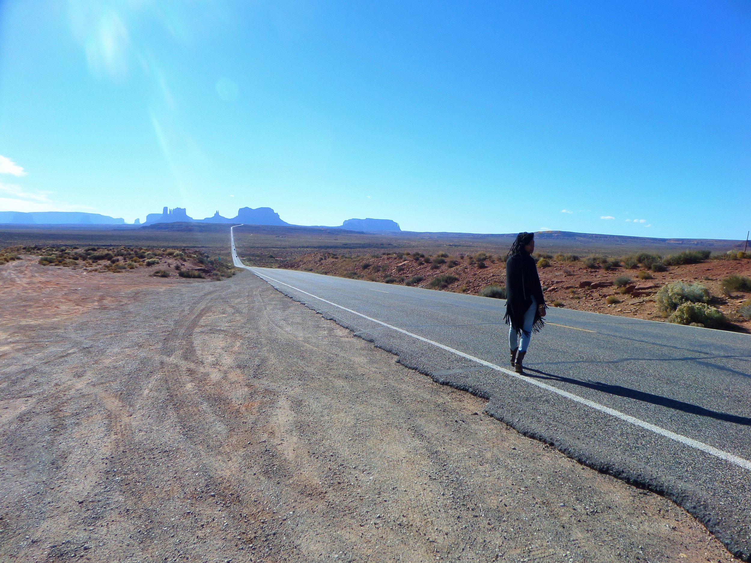Traveler beside a long desert highway disappearing into Monument Valley beneath bright Southwestern sunlight.