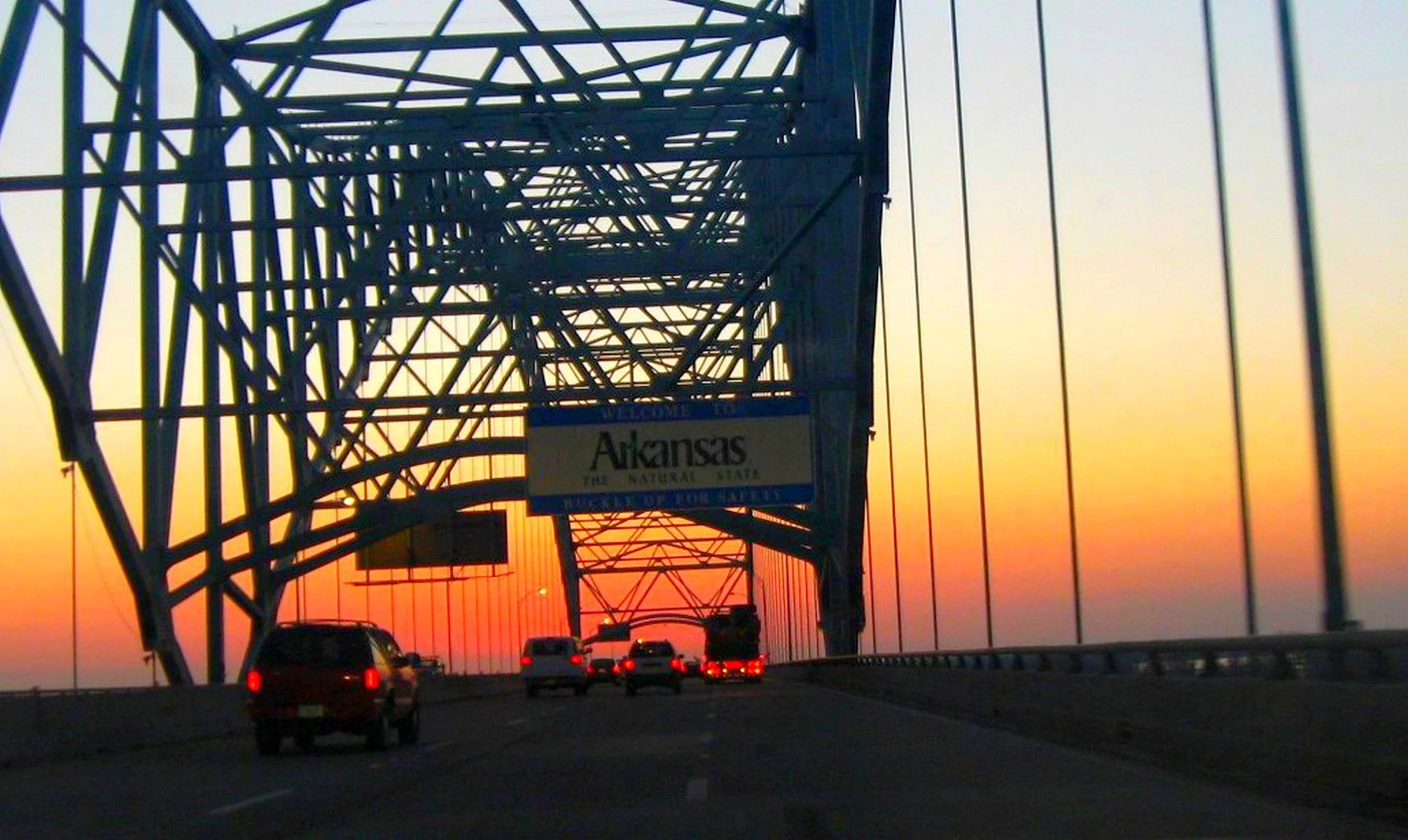 A highway bridge during sunset with cars traveling, steel structures, and a sign that reads 'Arkansas' in the foreground.