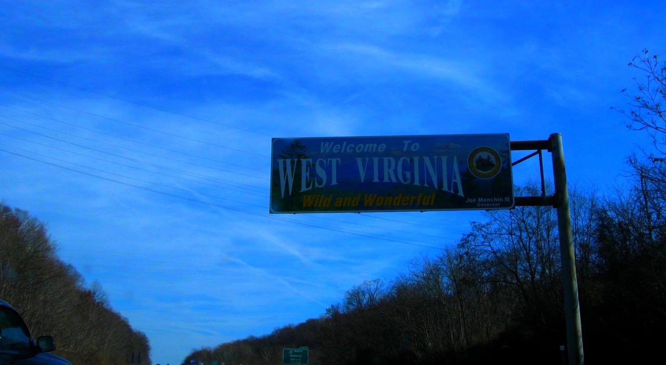 A highway sign welcoming travelers to West Virginia, with a slogan "Wild and Wonderful," under a blue sky with clouds and trees on either side of the road.
