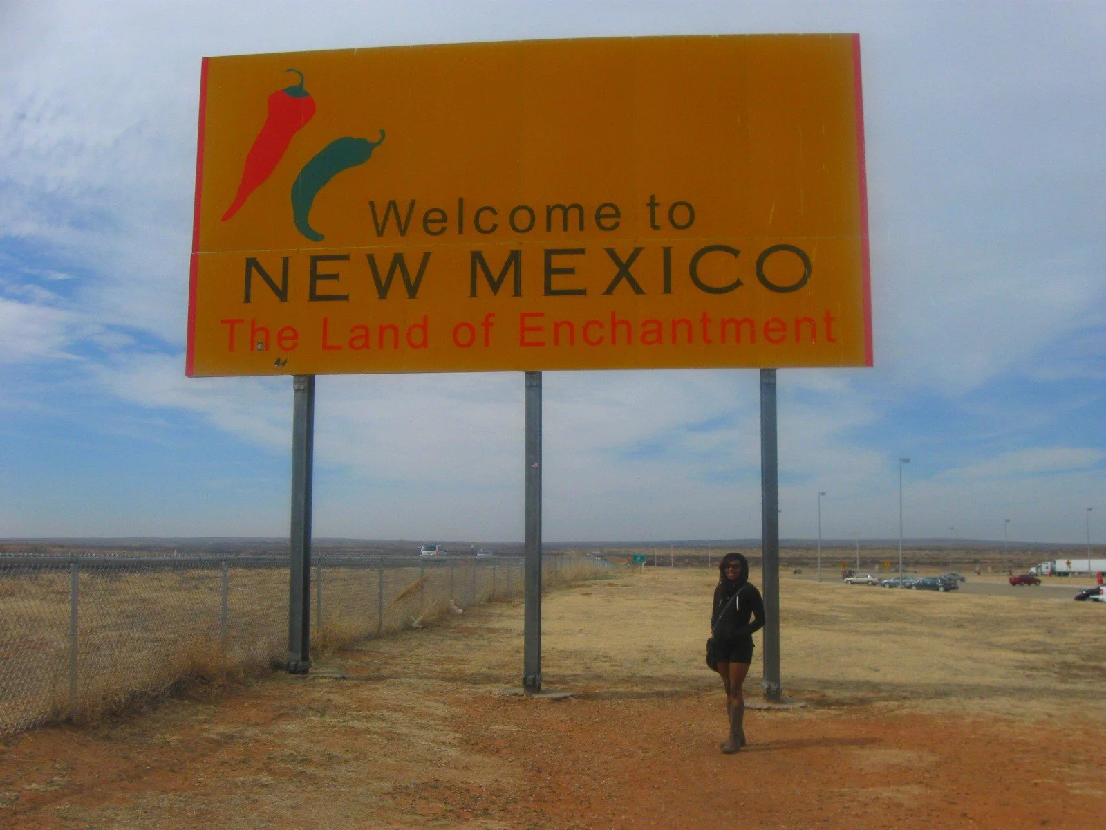 Large roadside sign welcoming visitors to New Mexico with chili pepper illustrations and the words "Welcome to New Mexico, The Land of Enchantment." A person stands near the sign on a dirt road, with a chain-link fence and a vast open landscape and h