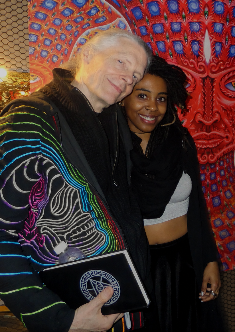 Bandit Belanger DaMori posing with visionary artist Alex Grey at the Gem & Jam Festival in Tucson, Arizona, in front of one of his colorful paintings.
