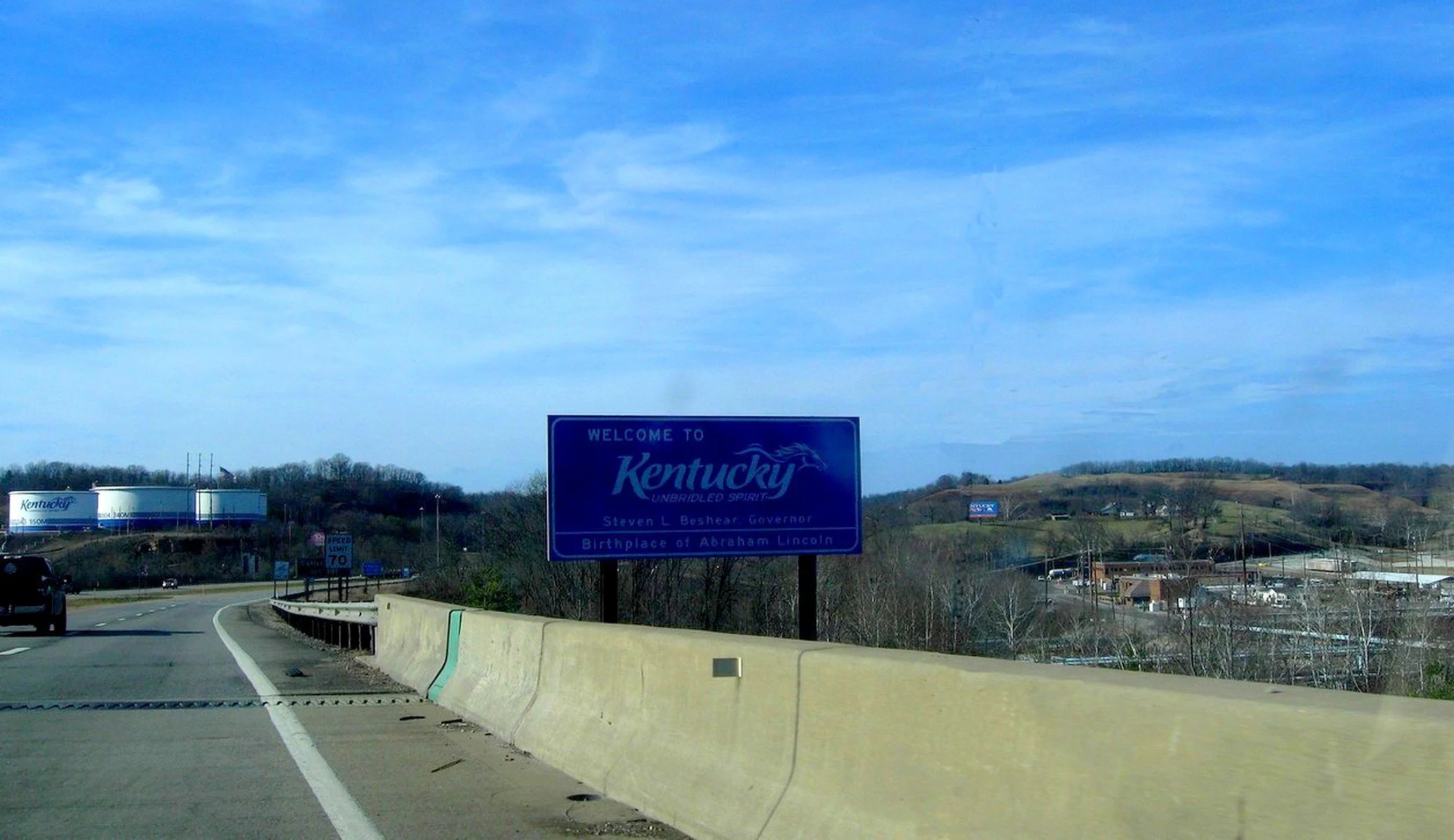 A highway leading into Kentucky with a large blue welcome sign reading "Welcome to Kentucky, Unbridled Spirit, Steven L. Beshear Governor, Birthplace of Abraham Lincoln." There are hills in the background and a billboard on the right side of the road