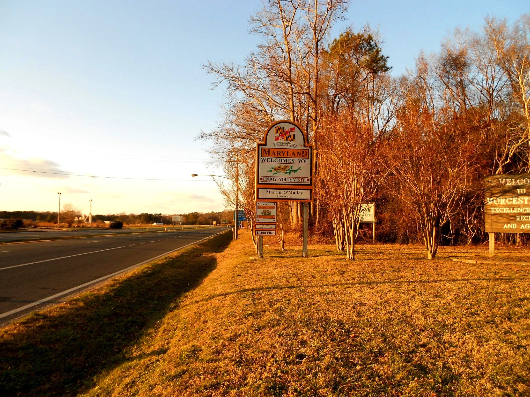 A roadside scene with a Maryland welcome sign and a row of leafless trees in the late afternoon sunlight. The sky is mostly clear with some clouds, and the road stretches into the distance.