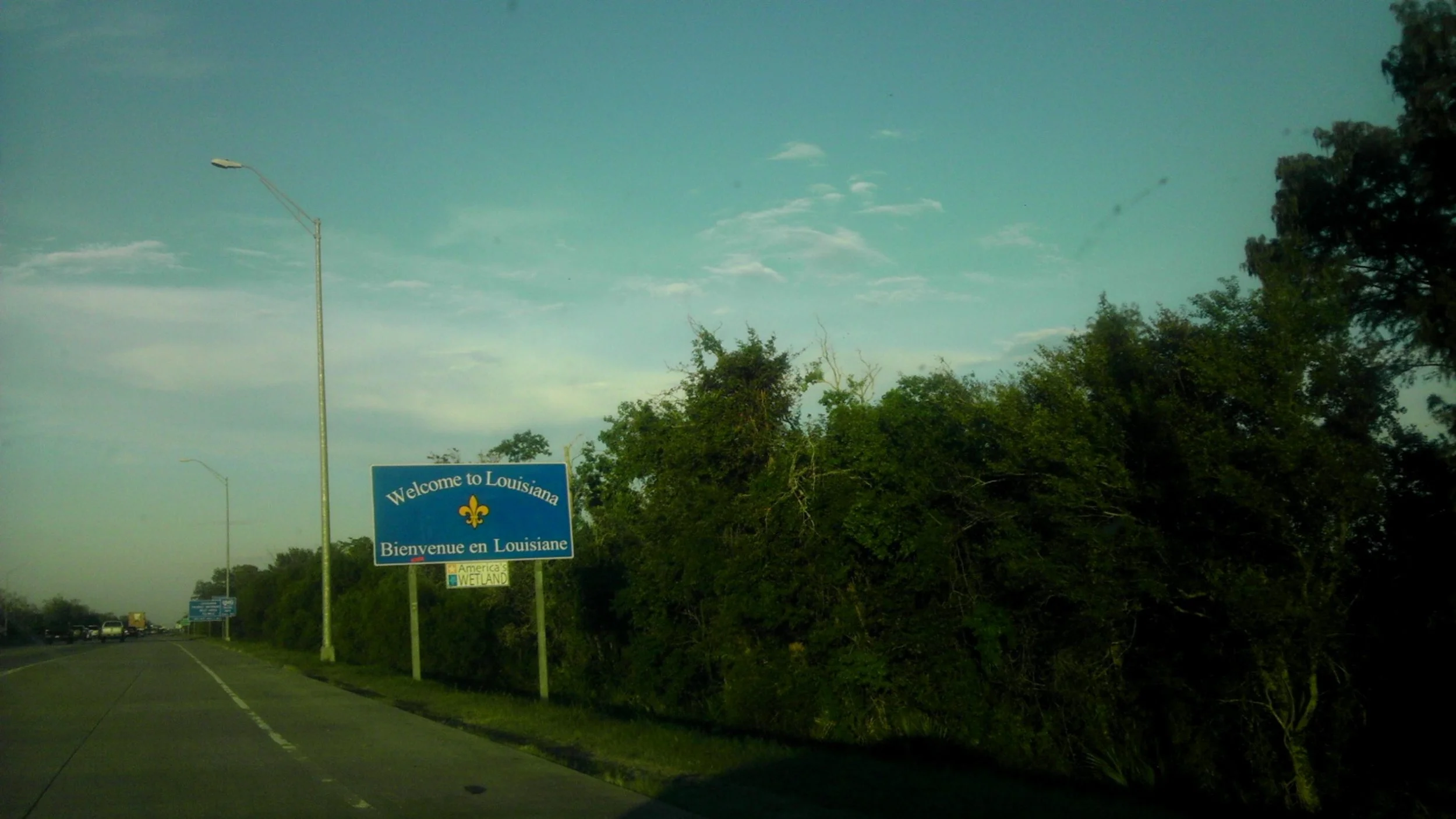 A roadside sign saying "Welcome to Louisiana" in English and French, with greenery and trees in the background under a partly cloudy sky.
