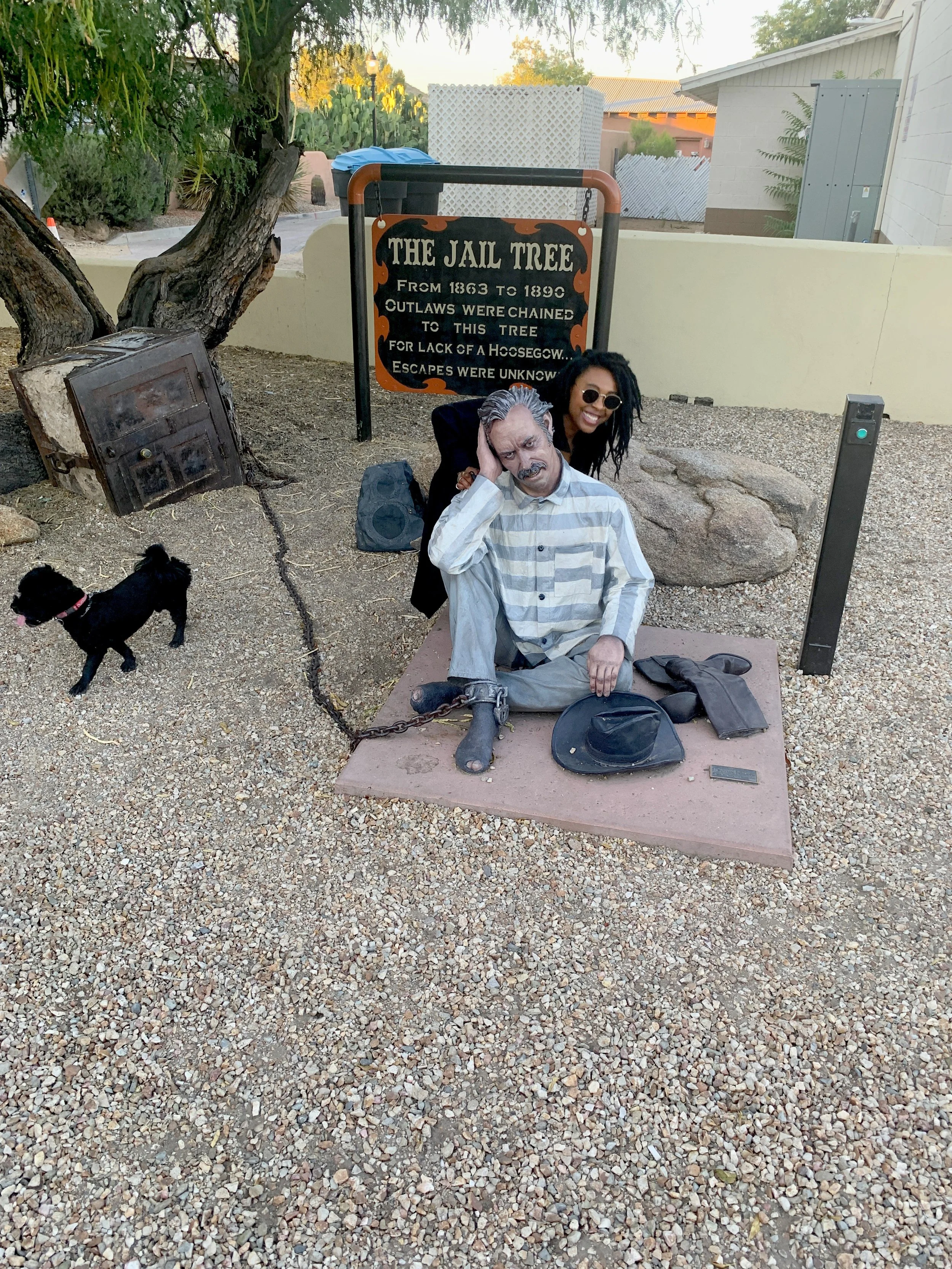 Woman with statue of an outlaw chained beneath the historic Wickenburg Jail Tree with interpretive sign in Wickenburg, Arizona.