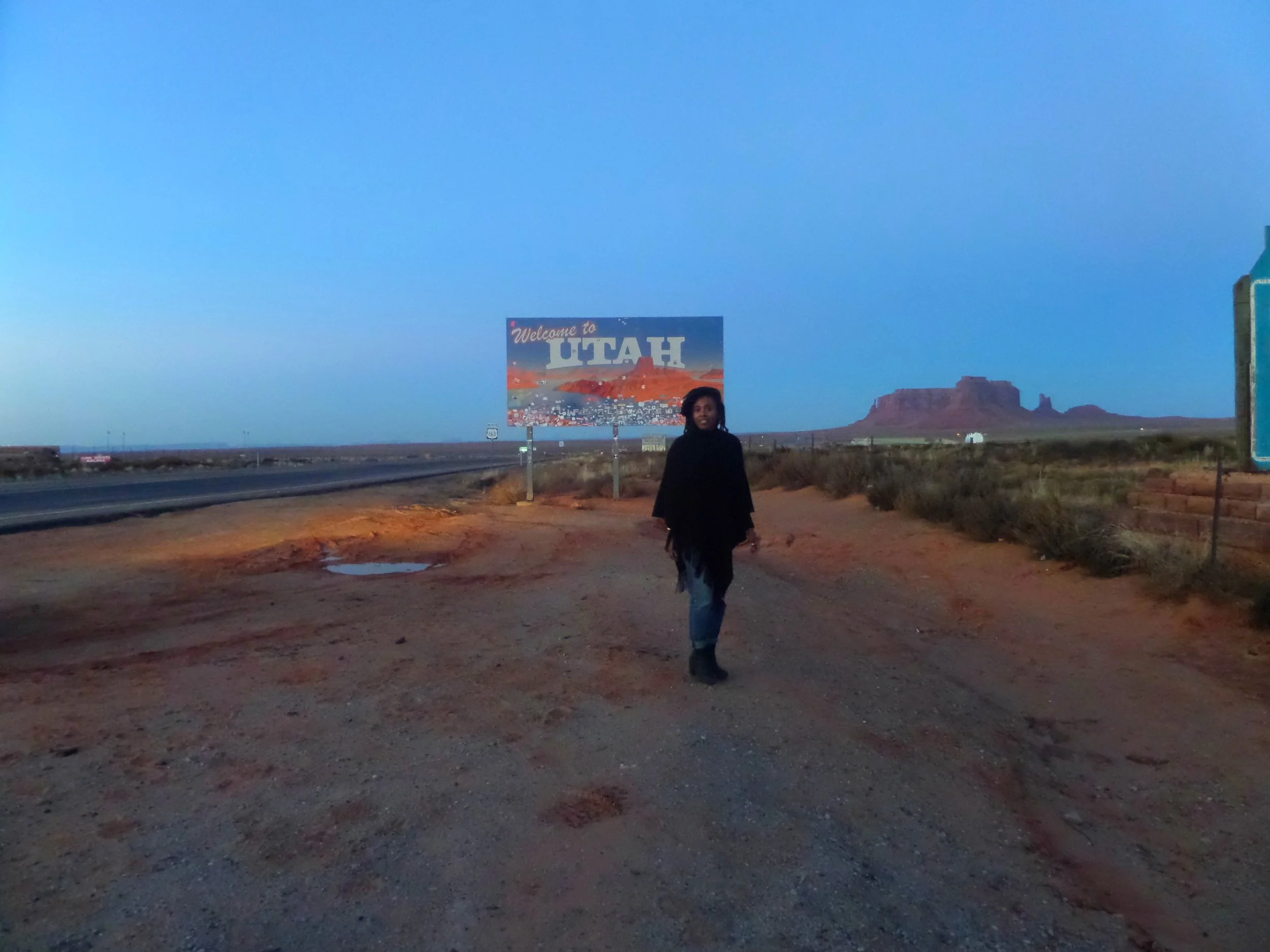 Person walking along a rural roadside at dusk with a welcome to Utah sign and scenic mountains in the background.