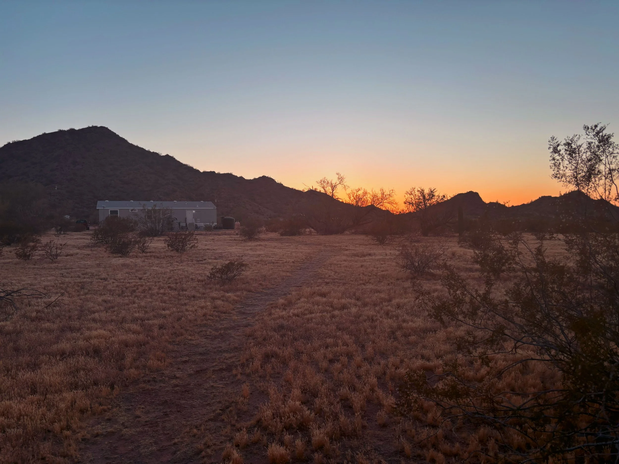 Desert landscape at sunset with a small modern house, sparse bushes, and mountainous terrain in the background.