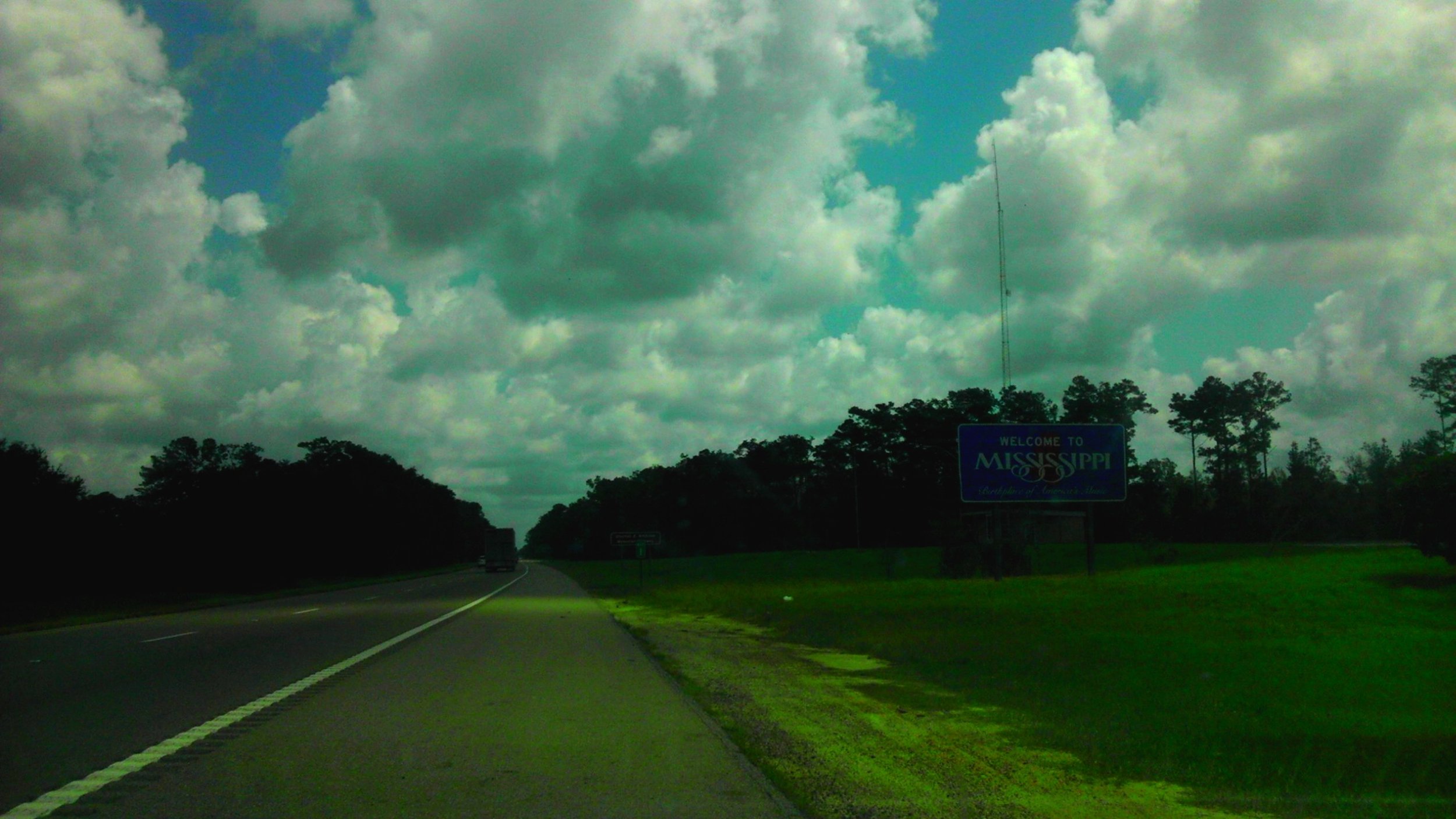 View of a highway with a sign that says 'Welcome to Mississippi' against a cloudy sky and trees lining the road.