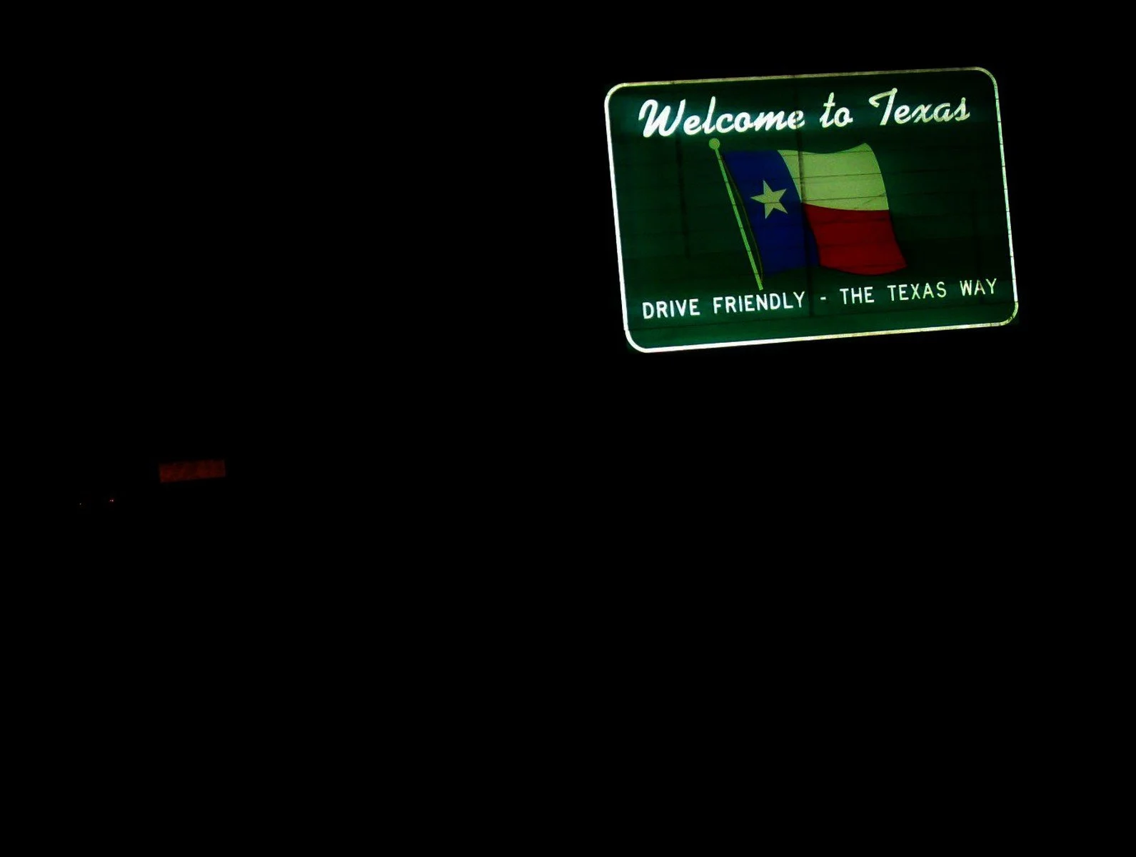 A neon-lit green roadside sign reads "Welcome to Texas" and features a Texas flag with a blue vertical stripe, a white star, and horizontal red and white stripes. The sign includes the message "Drive Friendly - The Texas Way" at the bottom.