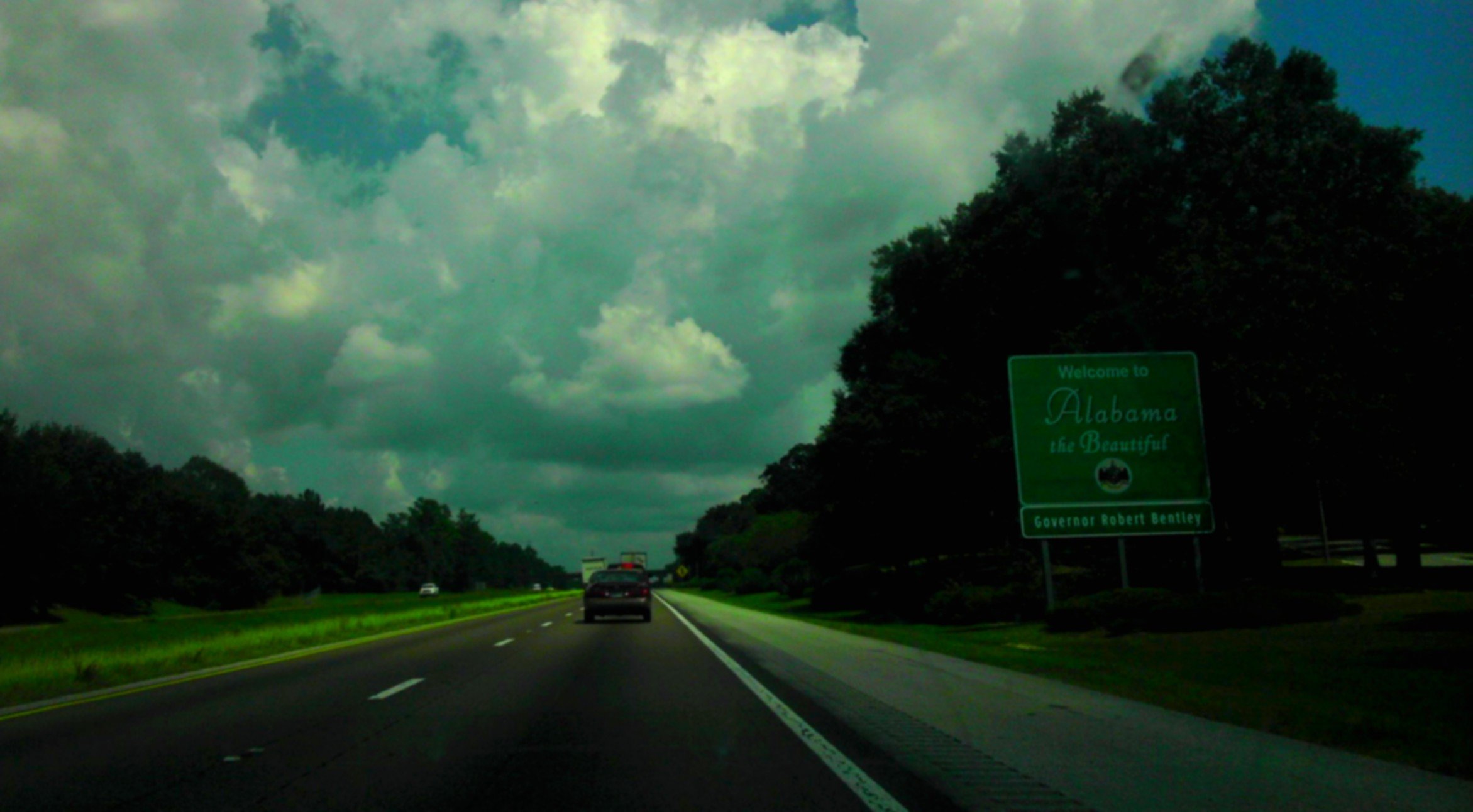 Highway driving into Alabama with a green welcome sign reading "Welcome to Alabama the Beautiful" and cloudy sky overhead.