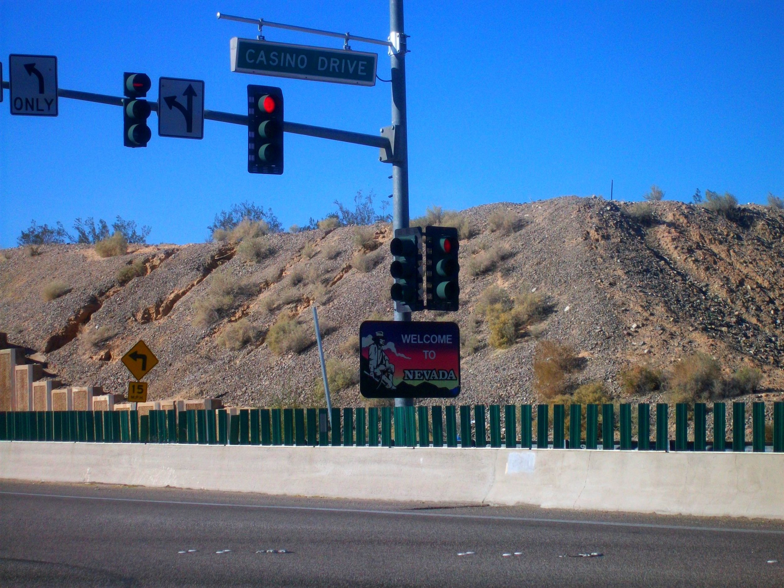 The image shows a roadside scene with traffic lights, road signs, and a welcome sign for Nevada. The background features rocky terrain and a clear blue sky.