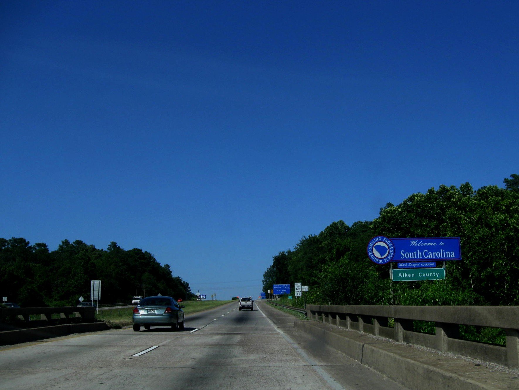 The image shows a highway with multiple cars driving towards the horizon under a mostly clear blue sky. There are green trees on both sides of the road and a blue sign indicating entry into South Carolina and Aiken County.