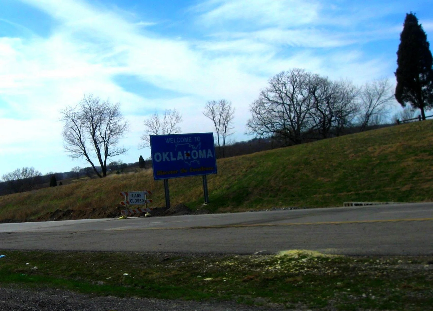 A blue welcome sign that says "Welcome to Oklahoma, Discover the Excellence" on the side of a road with leafless trees and a grassy hill in the background, and a smaller sign indicating a lane is closed.