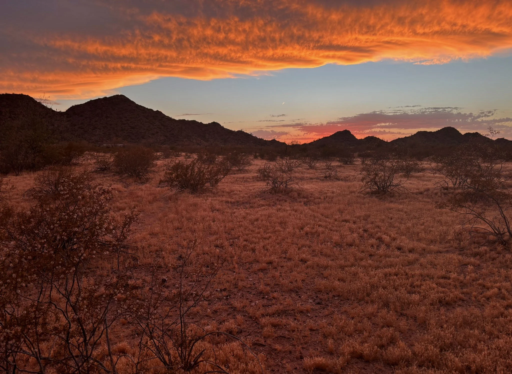 A desert landscape at sunset with orange and pink clouds, rocky hills in the distance, sparse desert bushes, and a dirt trail.