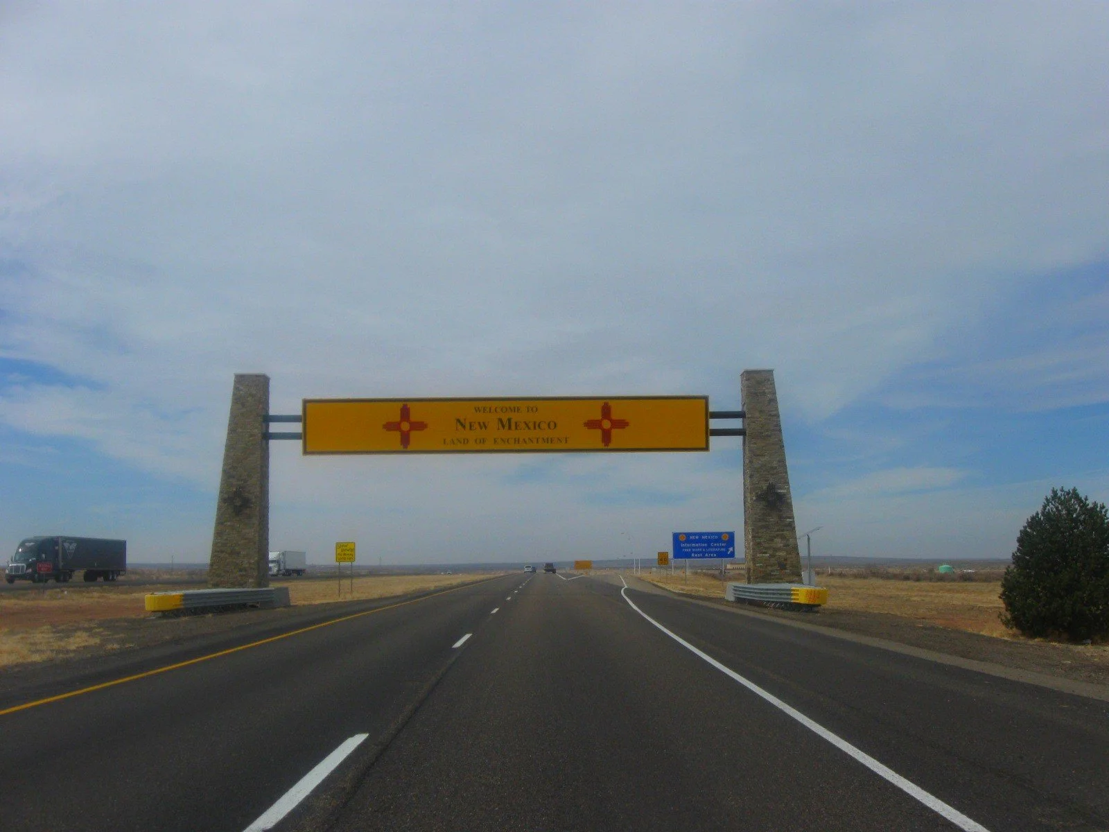 A highway sign welcoming travelers to New Mexico, labeled 'Land of Enchantment' with a yellow background and red and black text, supported by stone pillars on both sides of the road.