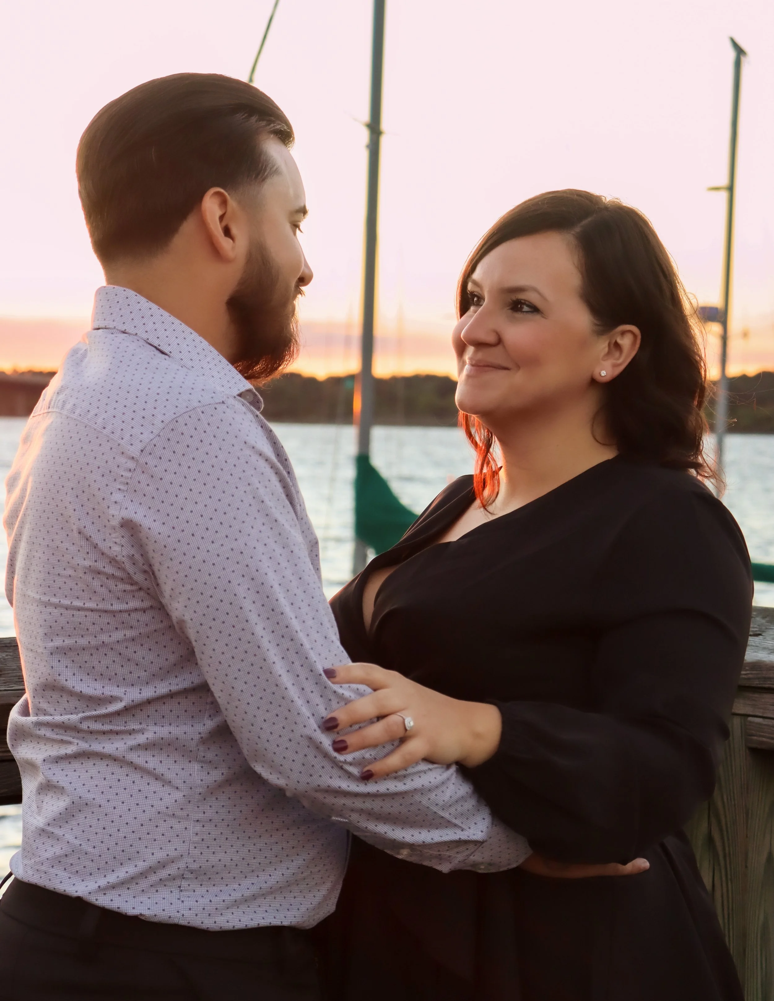 A couple standing close together outdoors near water at sunset, smiling at each other, with sailboats and a sunset in the background.