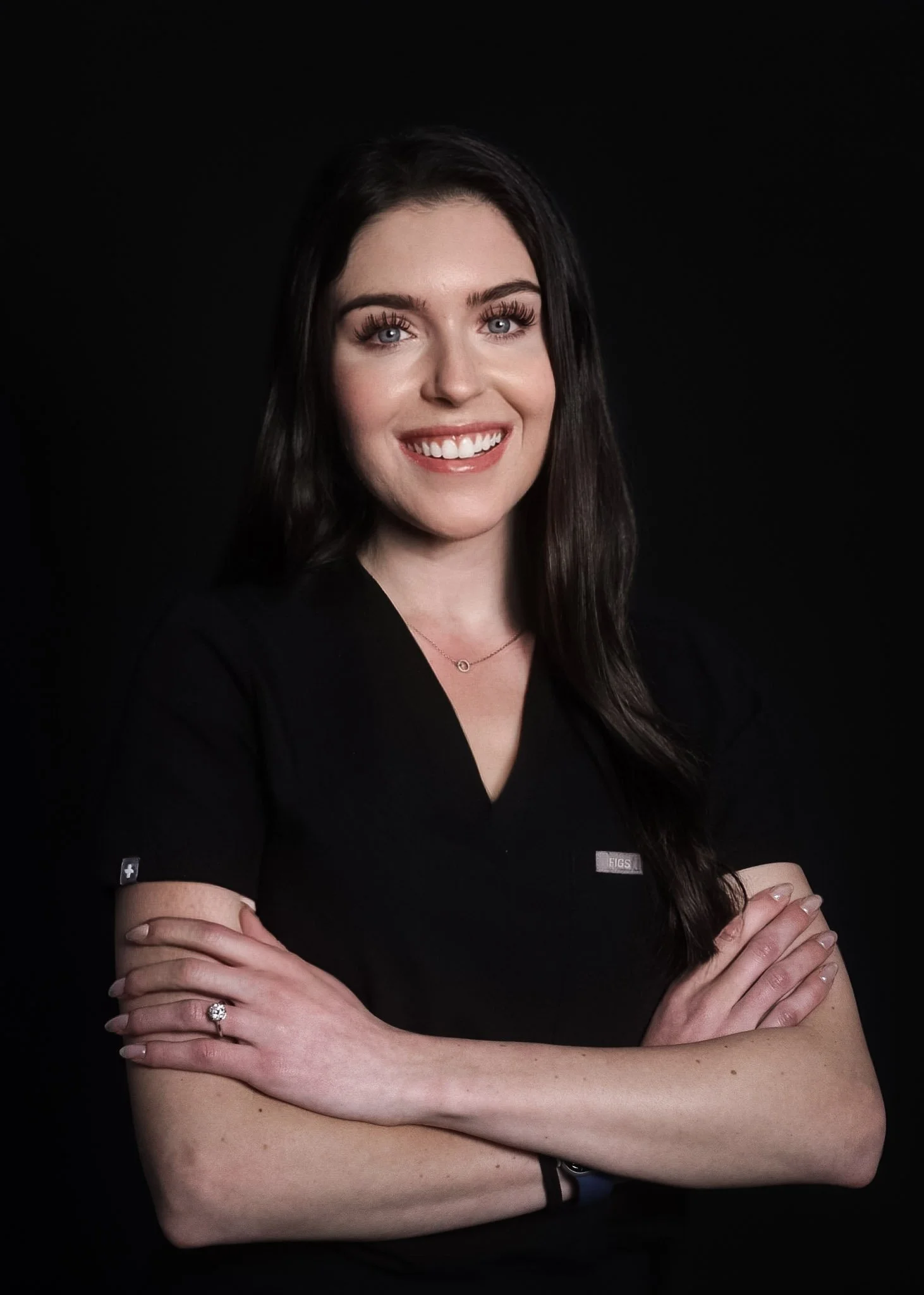 A smiling woman with long dark hair wearing a black V-neck top, crossed arms, and a delicate necklace against a black background.