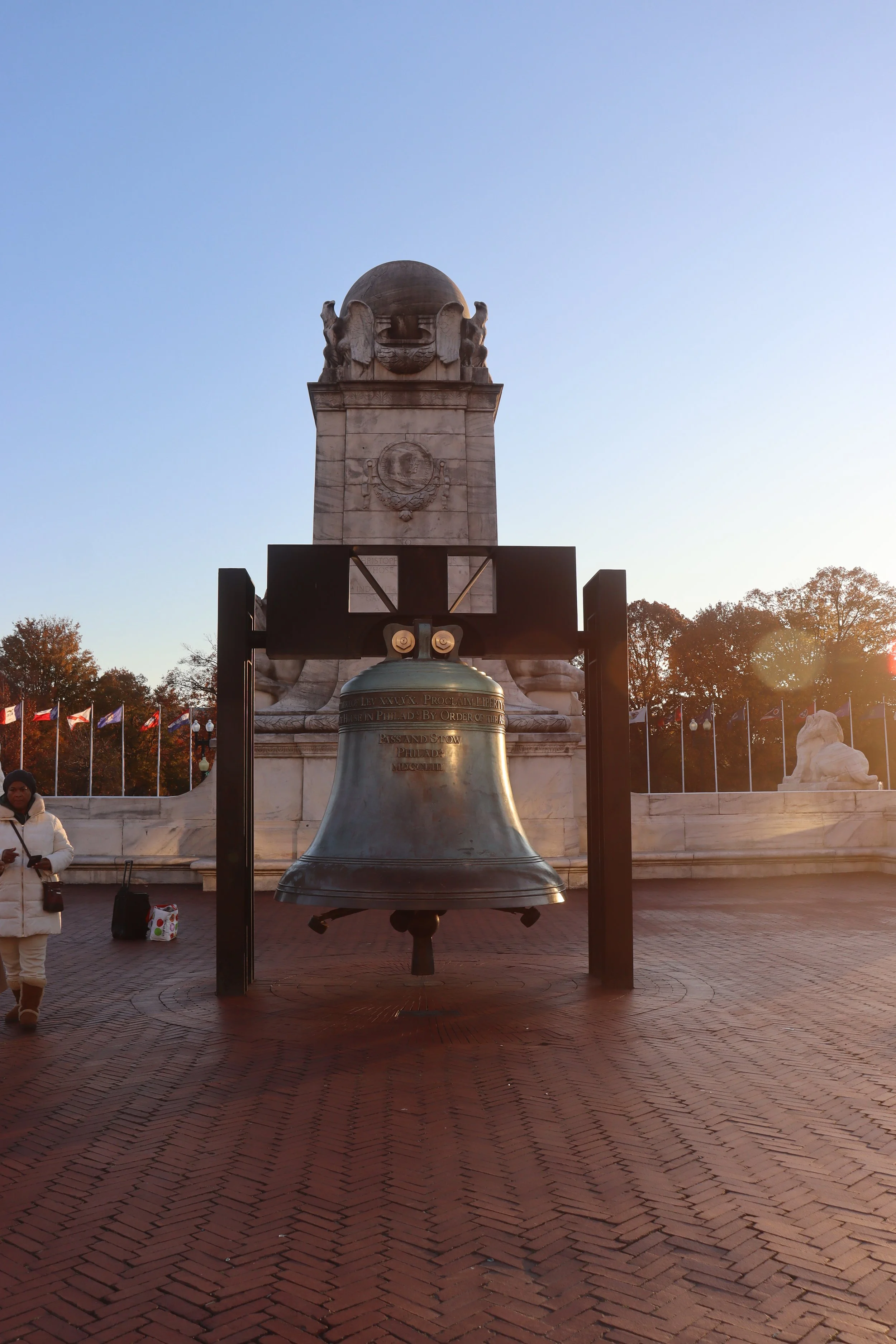 A large bell hanging in front of a stone monument with a globe and lion sculptures, located outdoors with flags in the background during sunset.