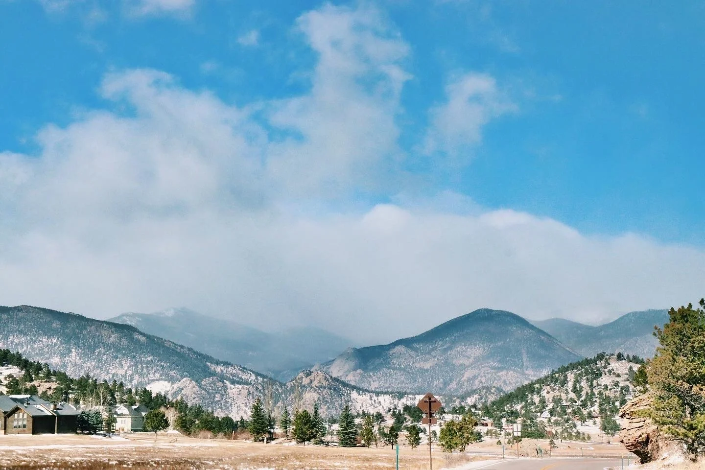Snow-capped mountains and trees with a partly cloudy blue sky, houses visible in the valley
