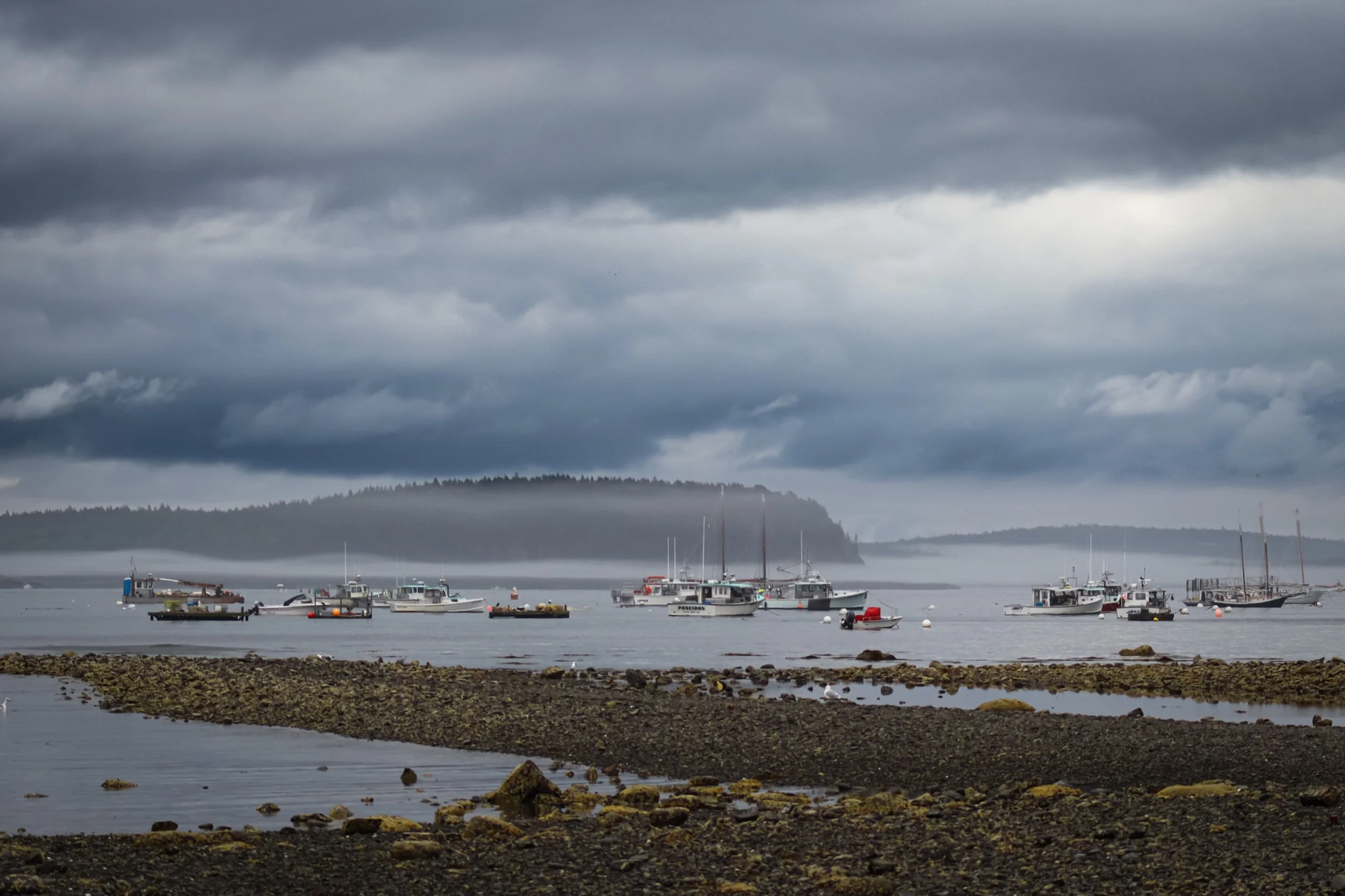 A cloudy sky over a calm harbor with several boats anchored near a rocky shoreline and fog-covered islands in the distance.