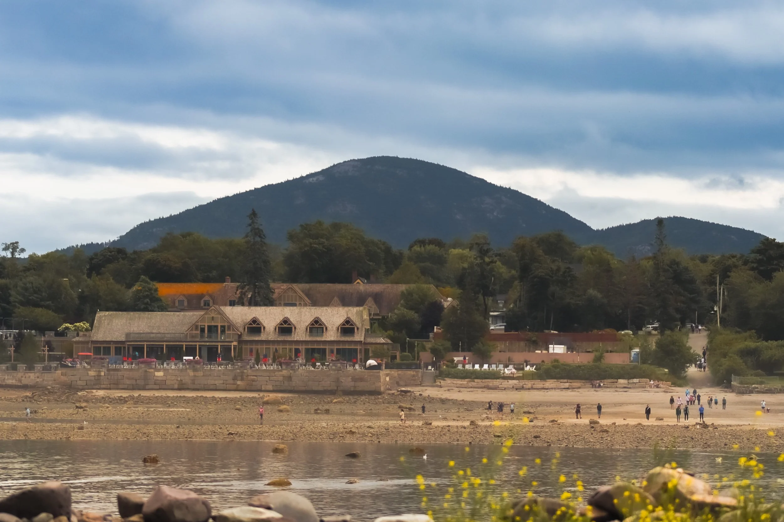 Scenic view of a lakeside with a building, trees, a mountain in the background, and people walking along the shoreline under a cloudy sky.
