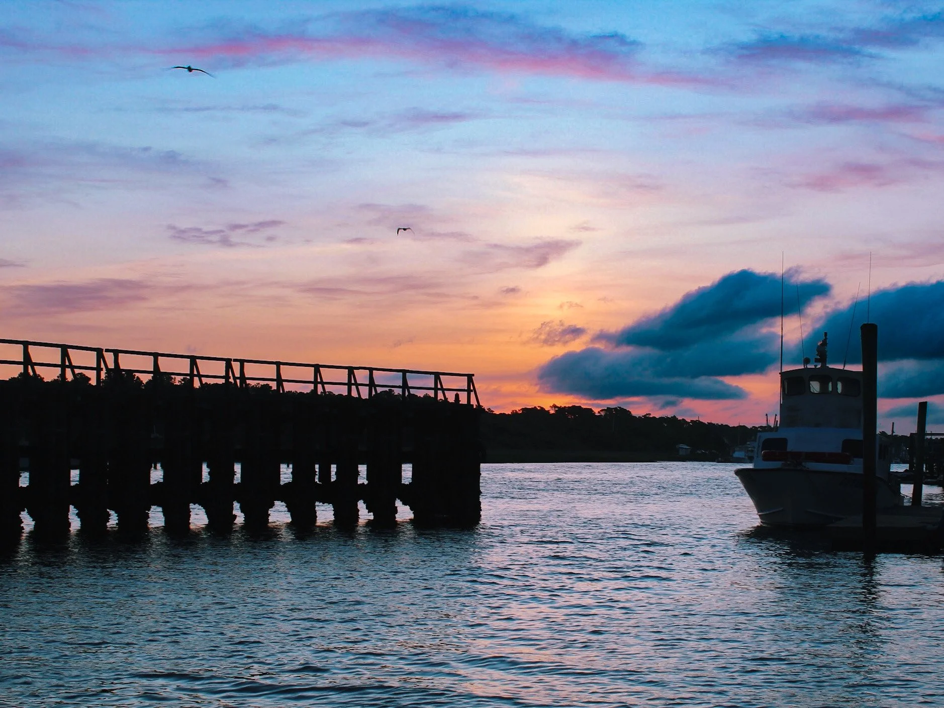 A sunset scene over a body of water with a wooden pier on the left and a boat on the right, with clouds and flying seagulls in the sky.