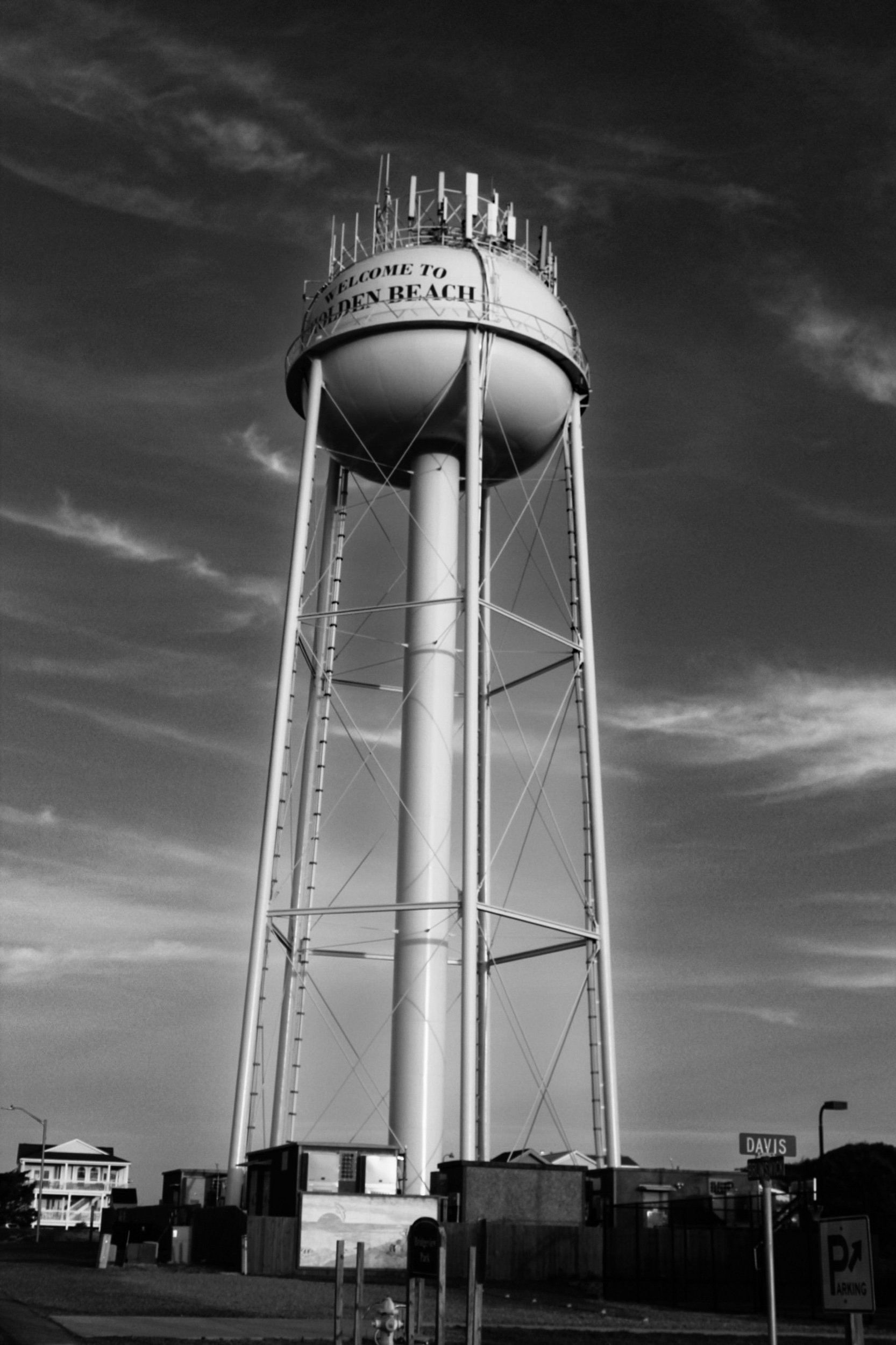 A tall water tower with a rounded top, featuring the words "Welcome to Golden Beach" on its side. The tower is surrounded by some buildings and street signs, including a sign for "Davis" and a parking sign. The sky is partly cloudy.