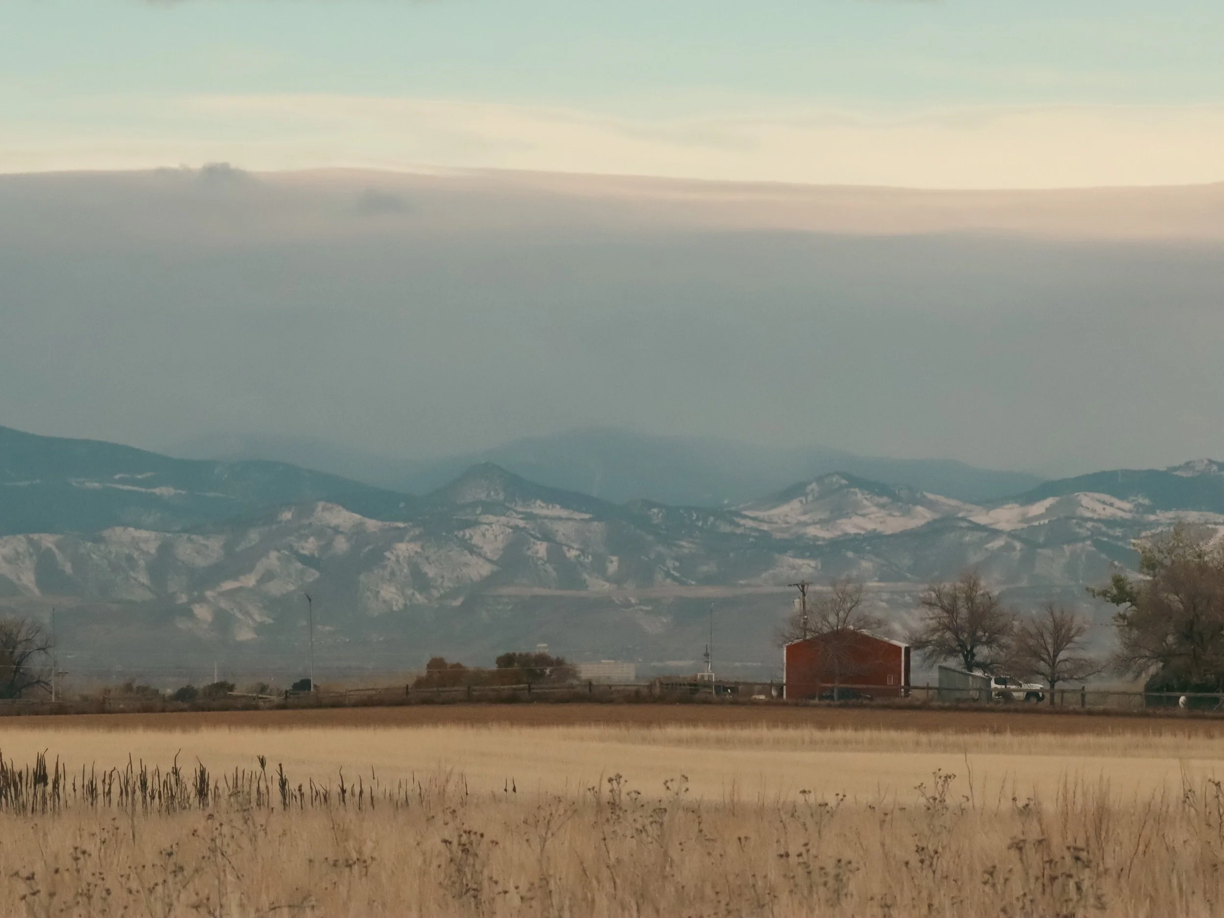 A landscape of snow-capped mountains under a partly cloudy sky, with a grassy field, trees, a small red barn, and utility poles in the foreground.