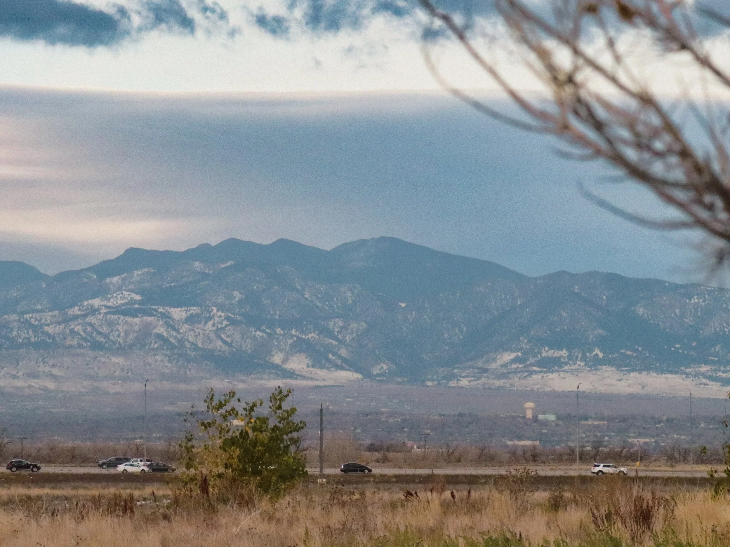 Scenic view of distant mountain range with snow-capped peaks, over a flat landscape with sparse vegetation and a highway with moving cars in the foreground.