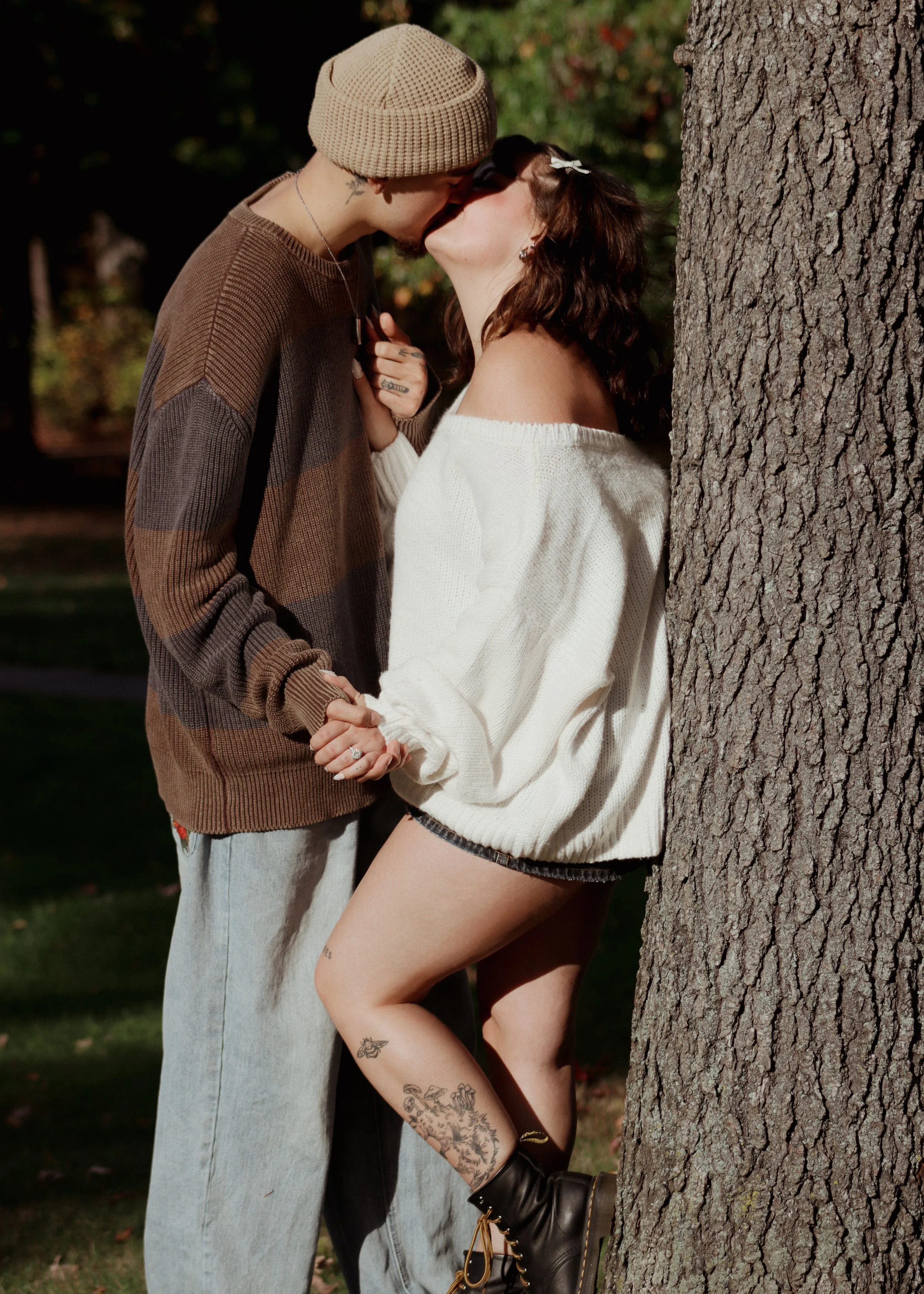 A couple kissing outdoors, holding hands, next to a tree with greenery in the background.