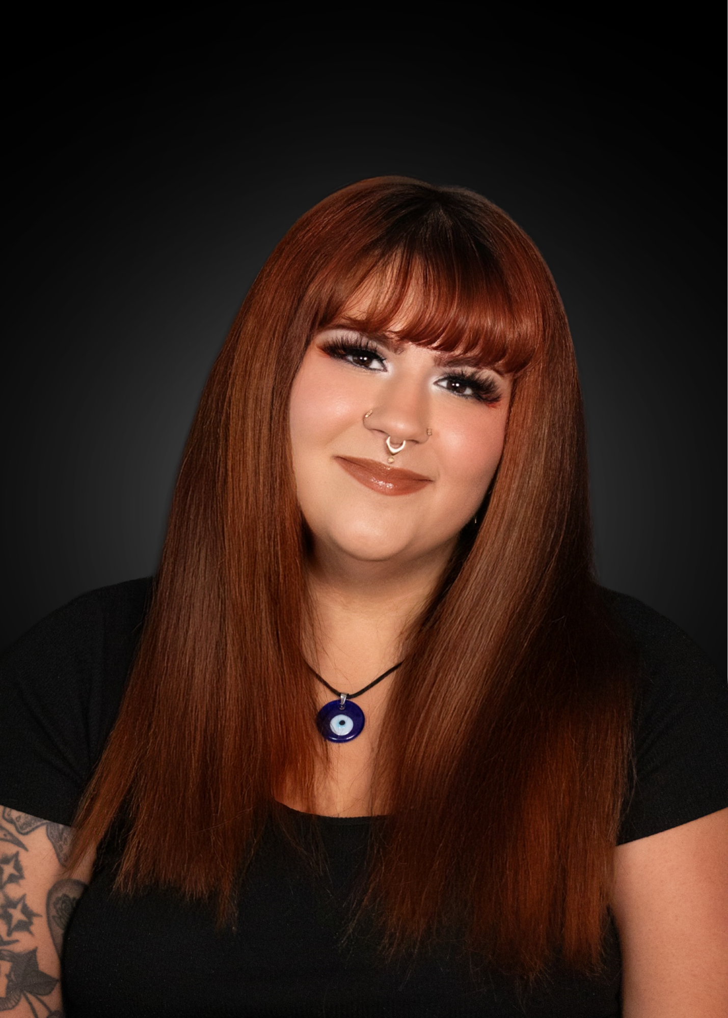 A woman with long, straight auburn hair and bangs, smiling against a dark gradient background. She has makeup, a nose ring, a septum piercing, and a necklace with a blue evil eye pendant.