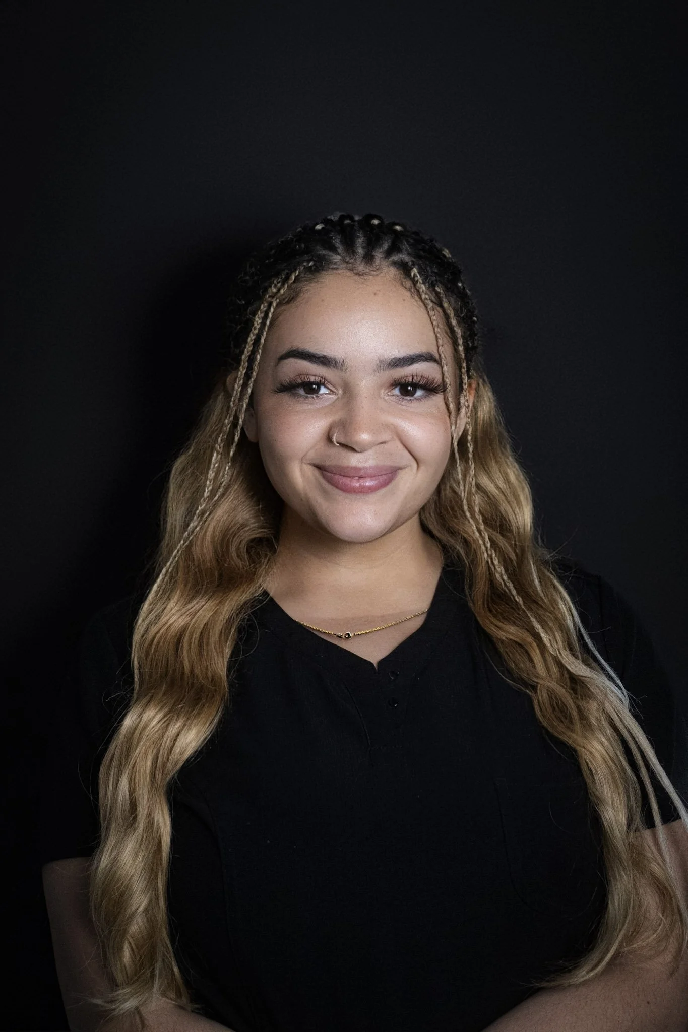 A young woman with long wavy blonde hair, braided on the top, smiling and wearing a black shirt against a dark background.