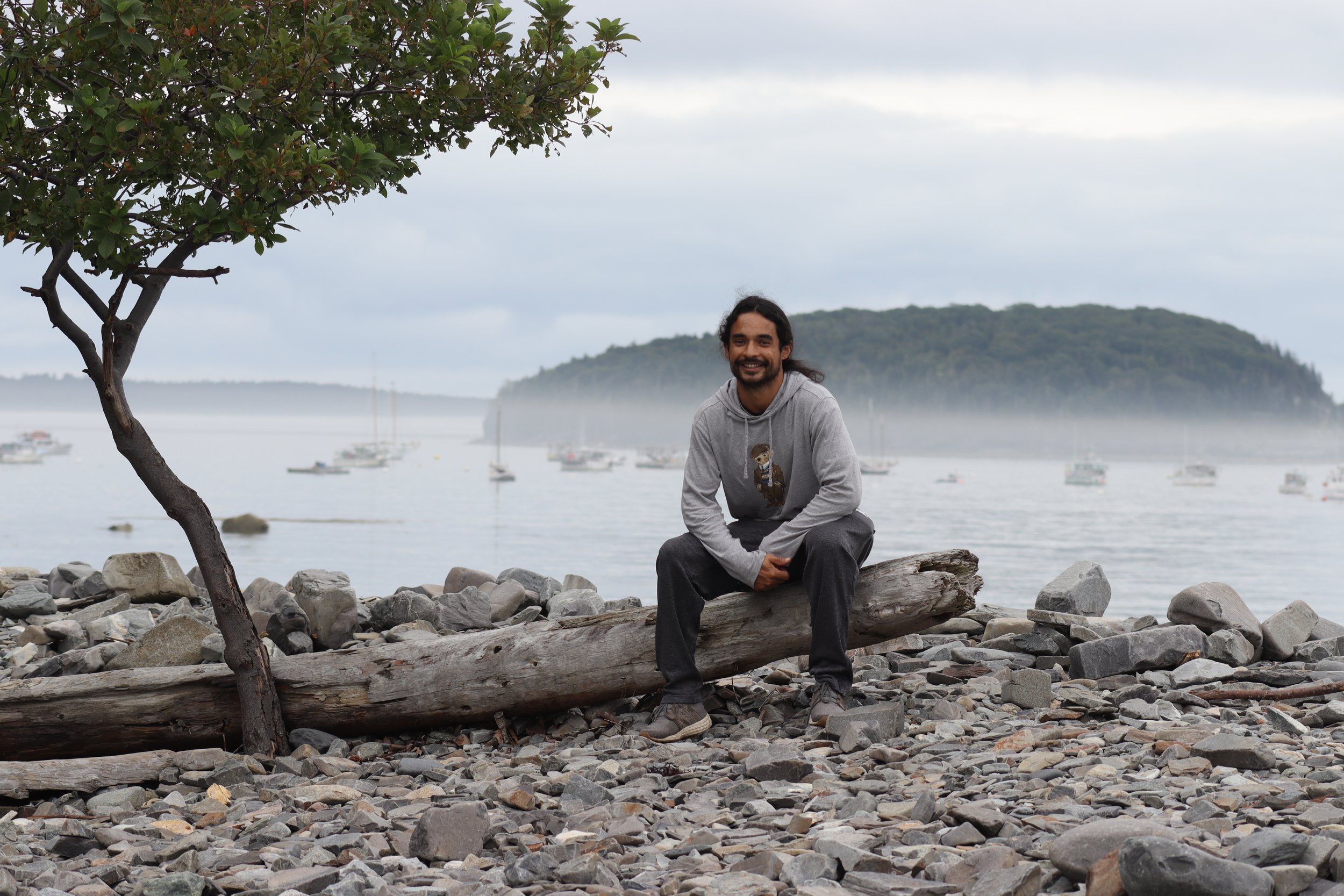 A man sitting on a driftwood log on a rocky beach with water, boats, and an island in the background. Overcast sky.