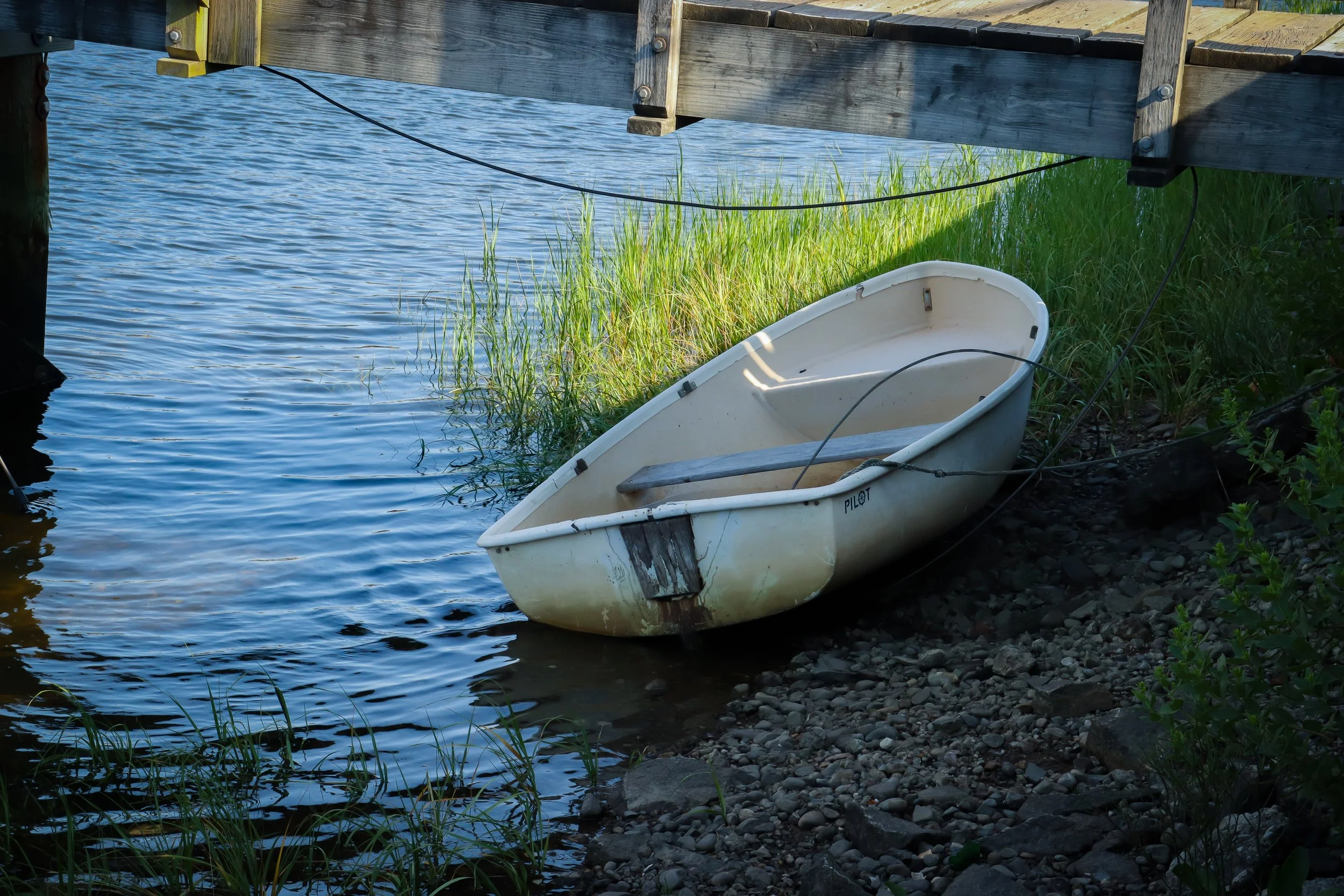 A small white rowboat resting on the rocky shoreline beneath a wooden dock near the water, tied with black ropes.