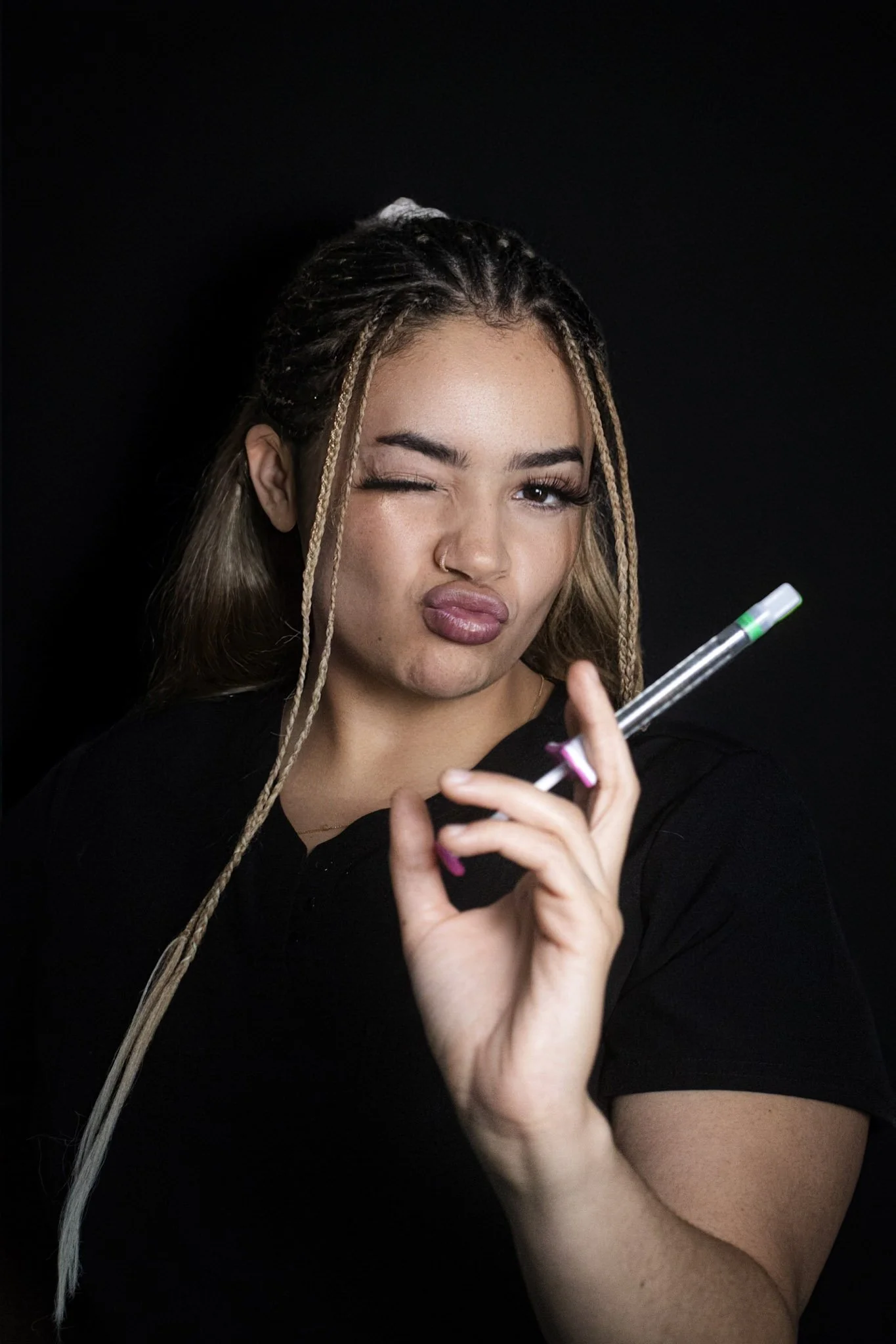 A woman with long hair styled in braids holding a clear test tube with a green cap, winking and making a pouty face against a black background.