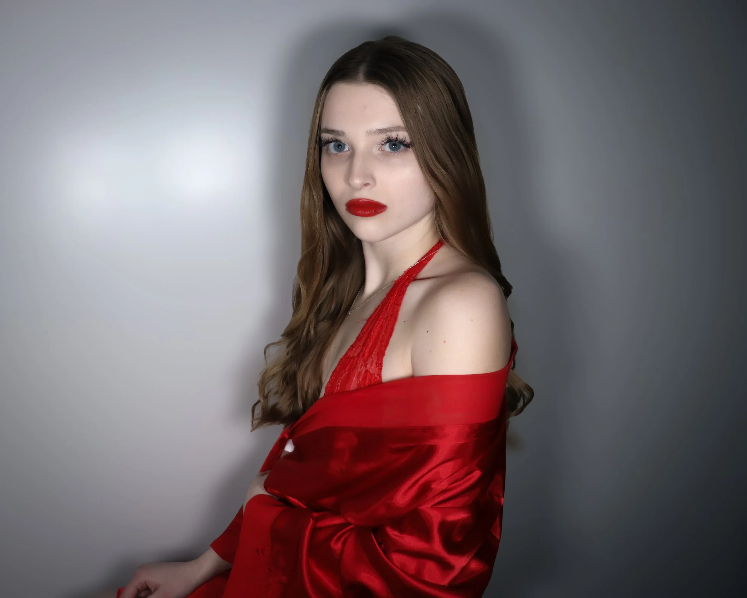A young woman with long brown hair and blue eyes wearing red lipstick, red satin robe, posing against a plain gray background.