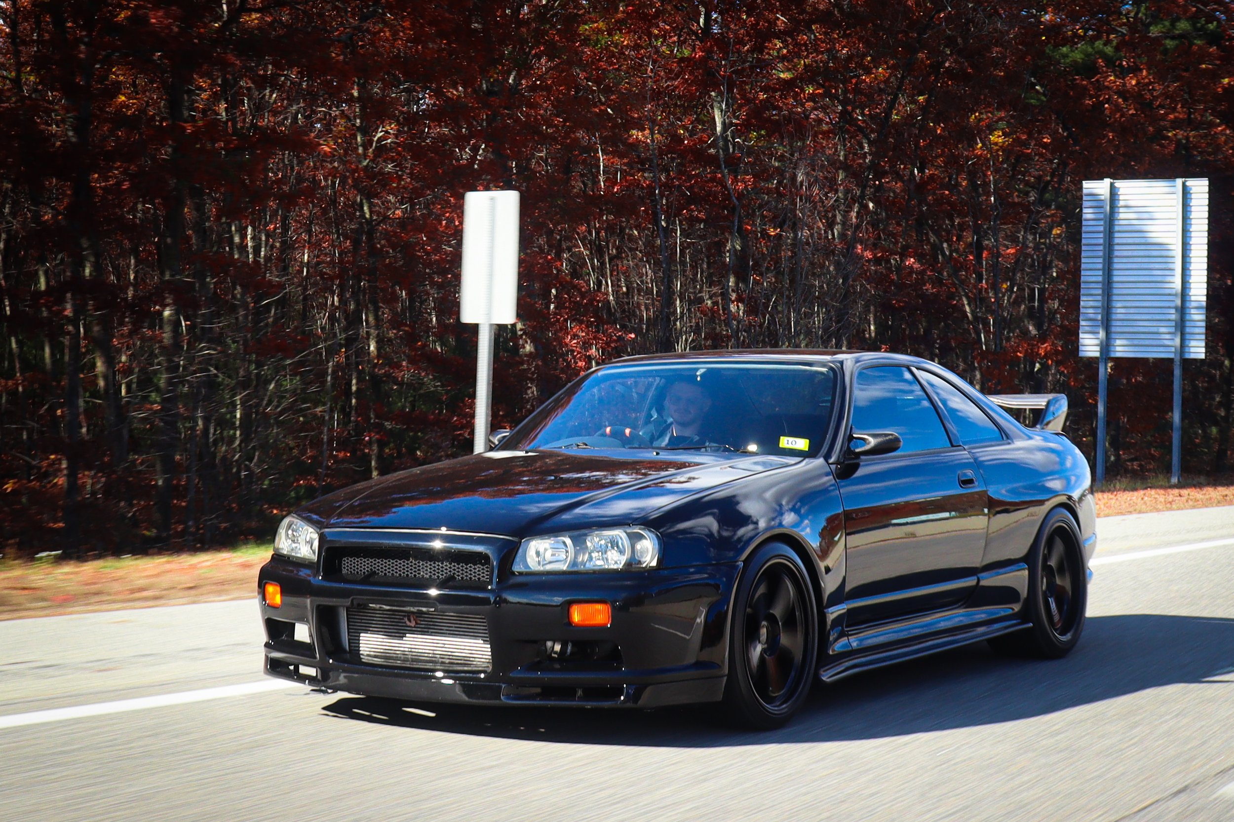 Black sports car driving on a highway with autumn trees in the background.