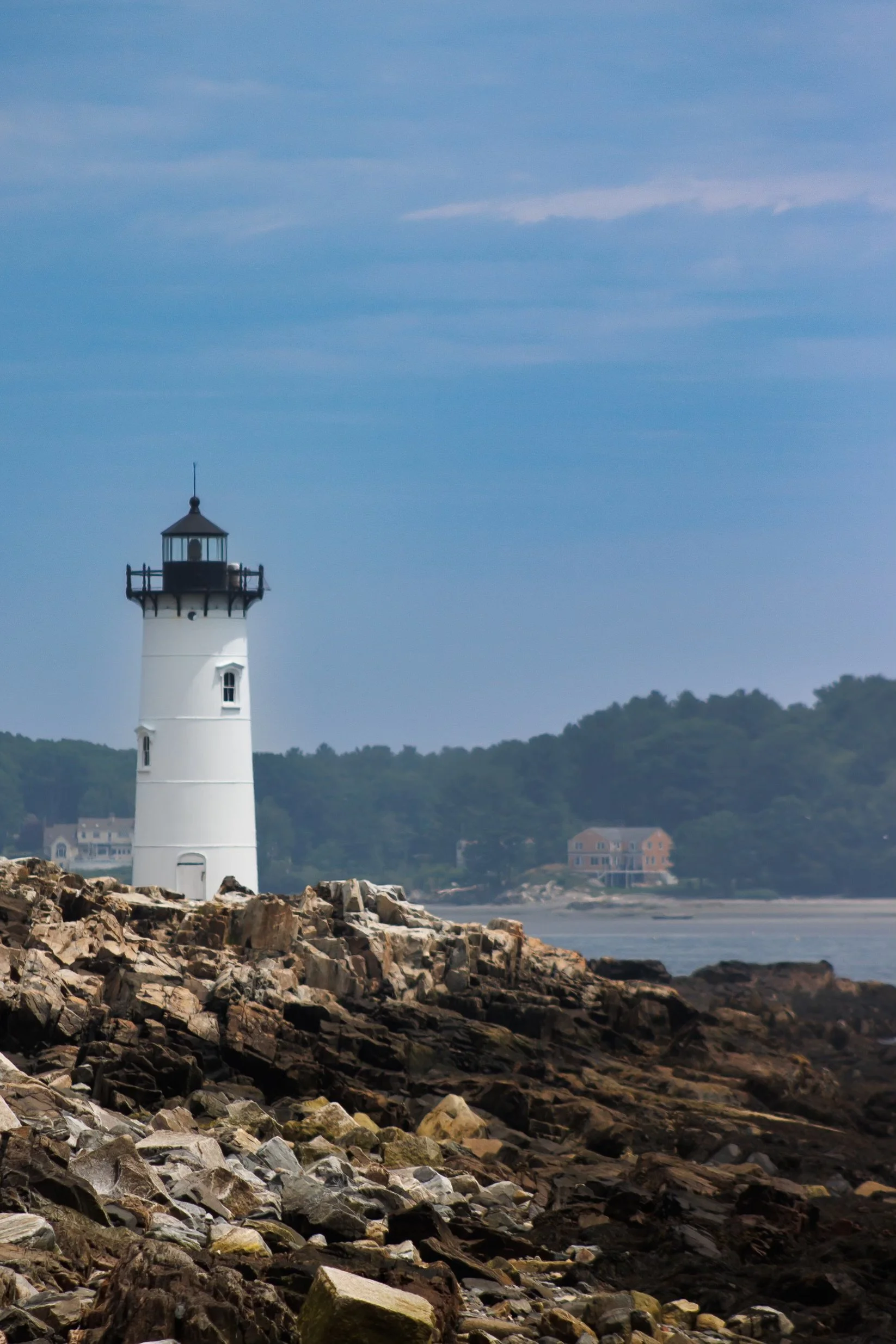 A lighthouse on rocky shoreline with water and distant houses in the background under a blue sky with wispy clouds.