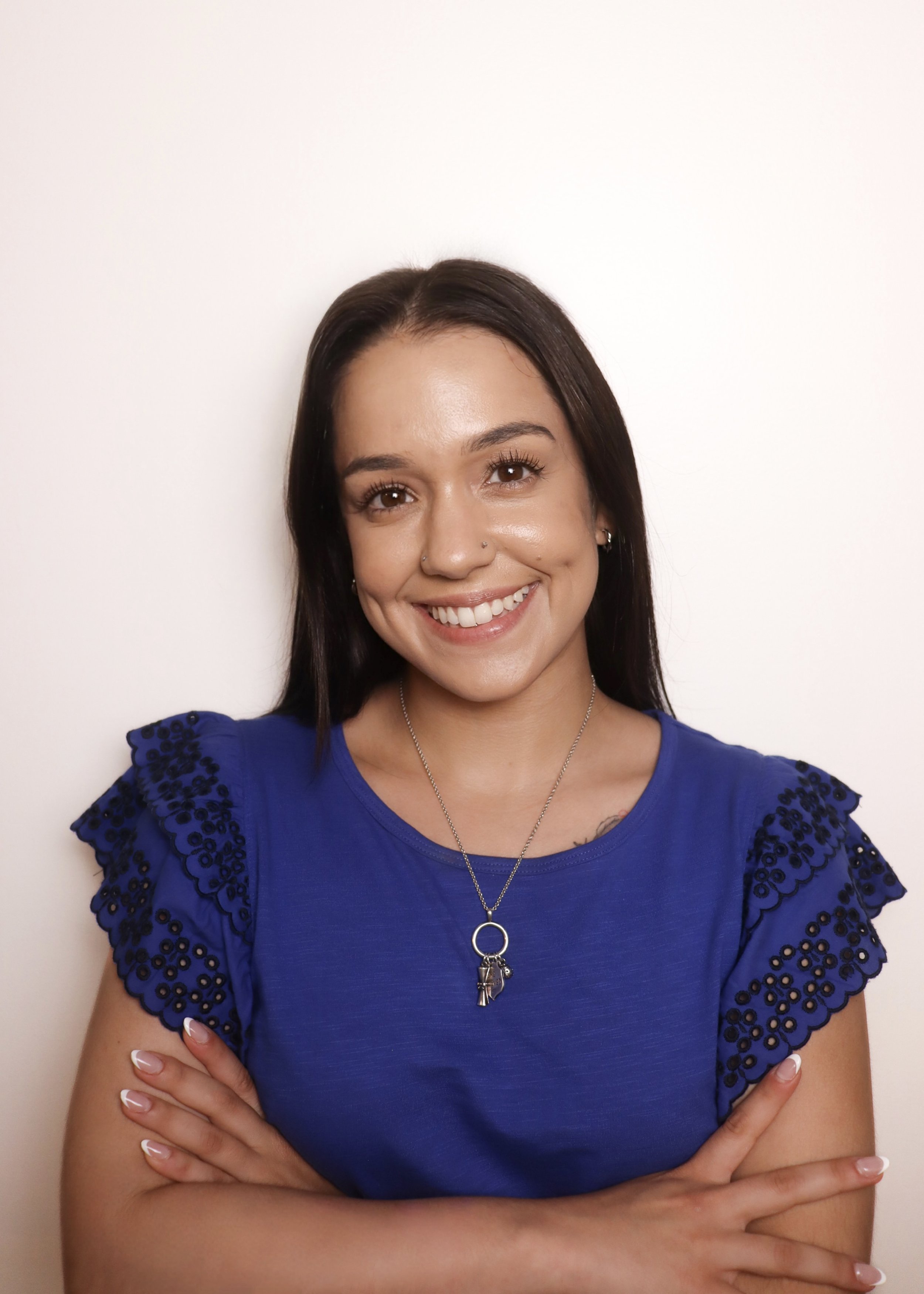 A young woman with long dark hair, wearing a blue top with lace details on the sleeves, smiling with arms crossed against a plain white background.