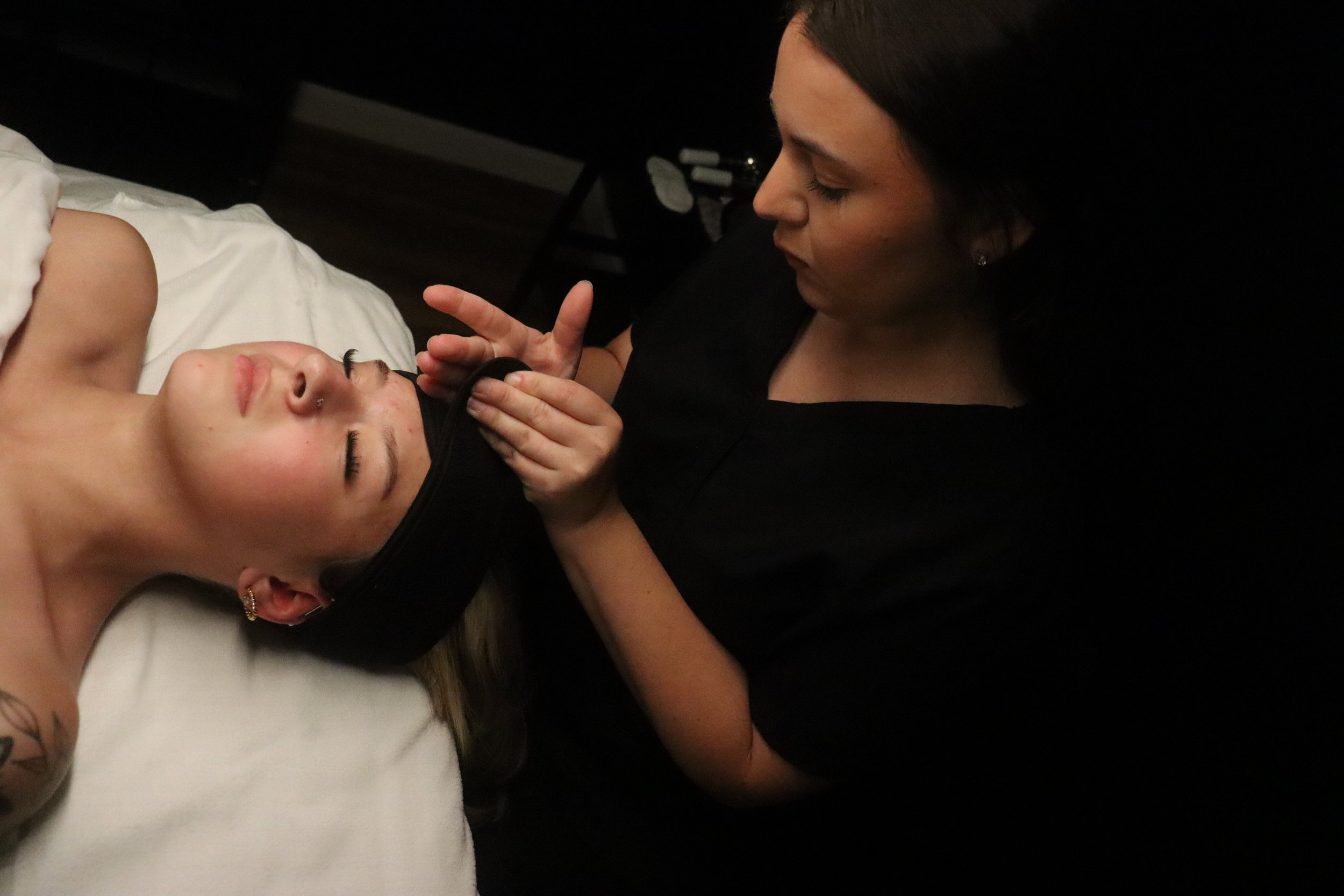 A woman is tending to a young girl lying on a bed. The girl has her eyes closed and is wearing a black headband. The woman appears to be adjusting or fixing the girl’s face or hair.