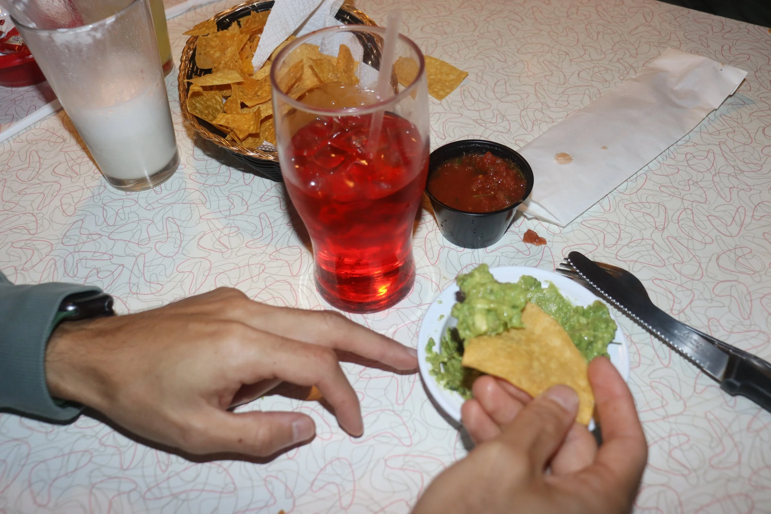 Person holding a small plate with guacamole and a tortilla chip, sitting at a table with a tall glass of iced red drink, a glass of milk, a bowl of salsa, and a basket of tortilla chips.