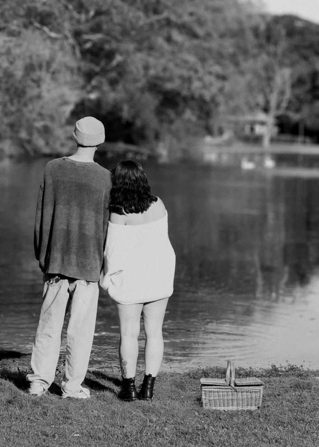 A black-and-white photo of a couple standing by a lake with trees in the background. The man is wearing a beanie and a sweater, and the woman is wrapped in a blanket or large scarf, both facing the water. There is a picnic basket on the grass nearby.