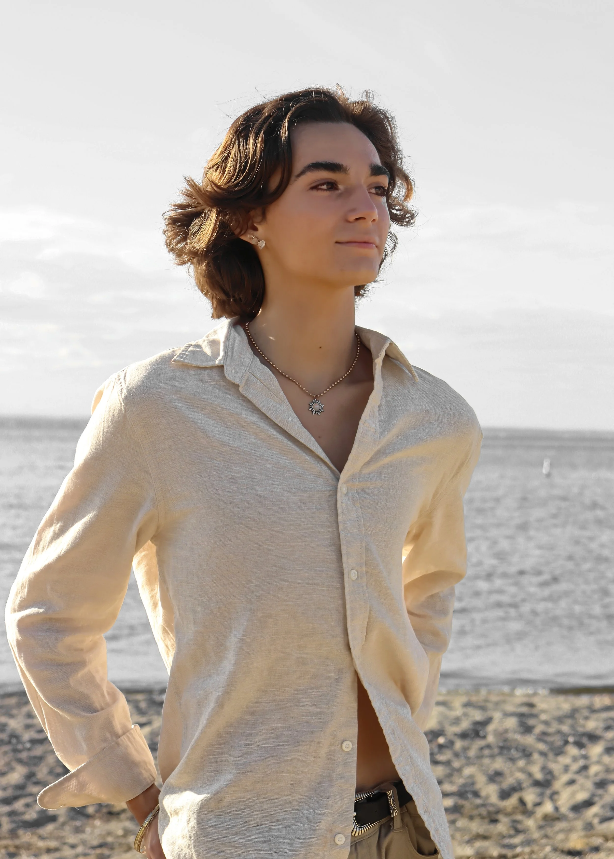 A young woman with short brown hair and light skin stands on a beach, wearing a beige button-up shirt, jewelry, and looking to the side. The background features the sea and sky.