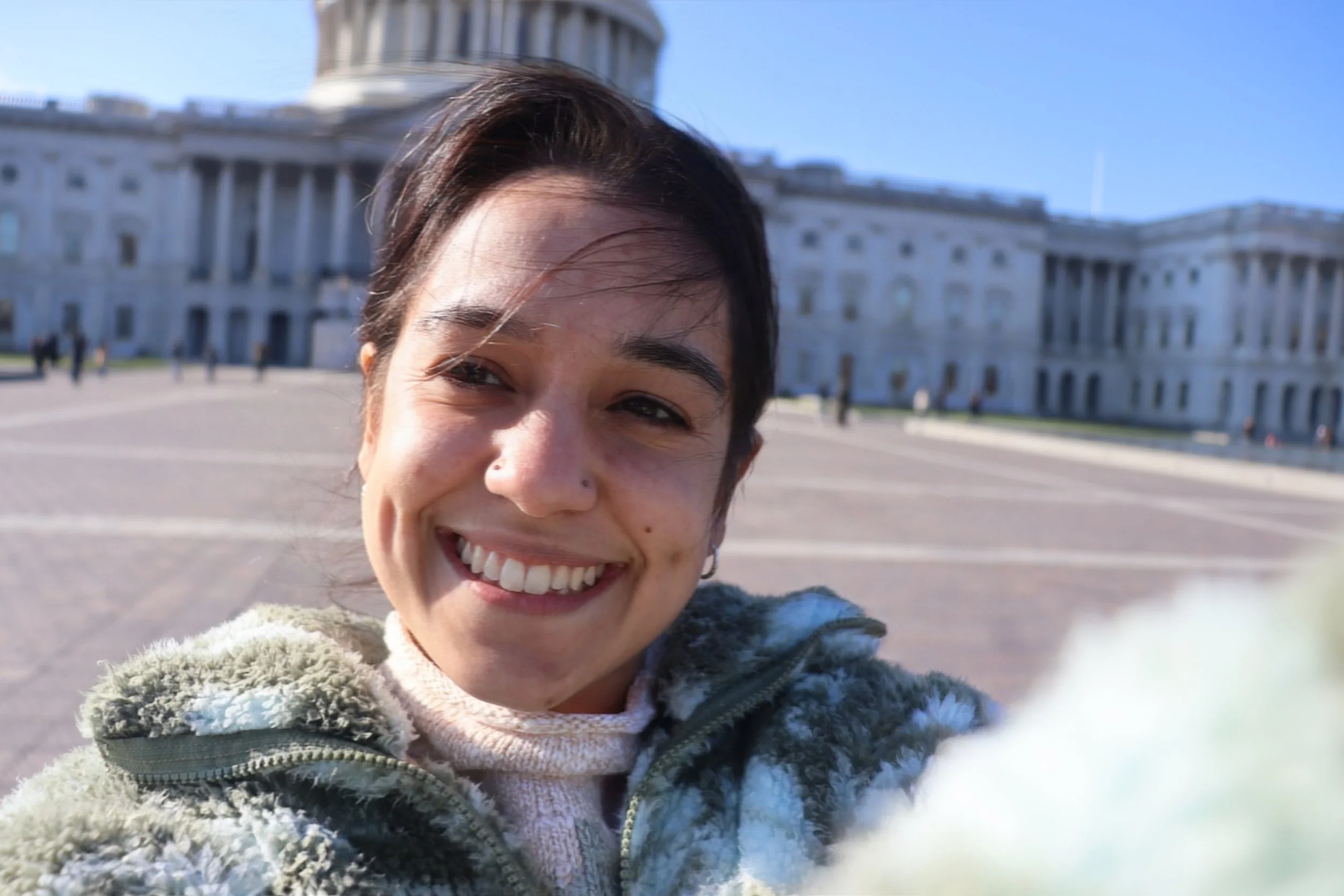 Close-up selfie of a smiling woman with short dark hair, wearing a fluffy green jacket, in front of the U.S. Capitol building on a sunny day.