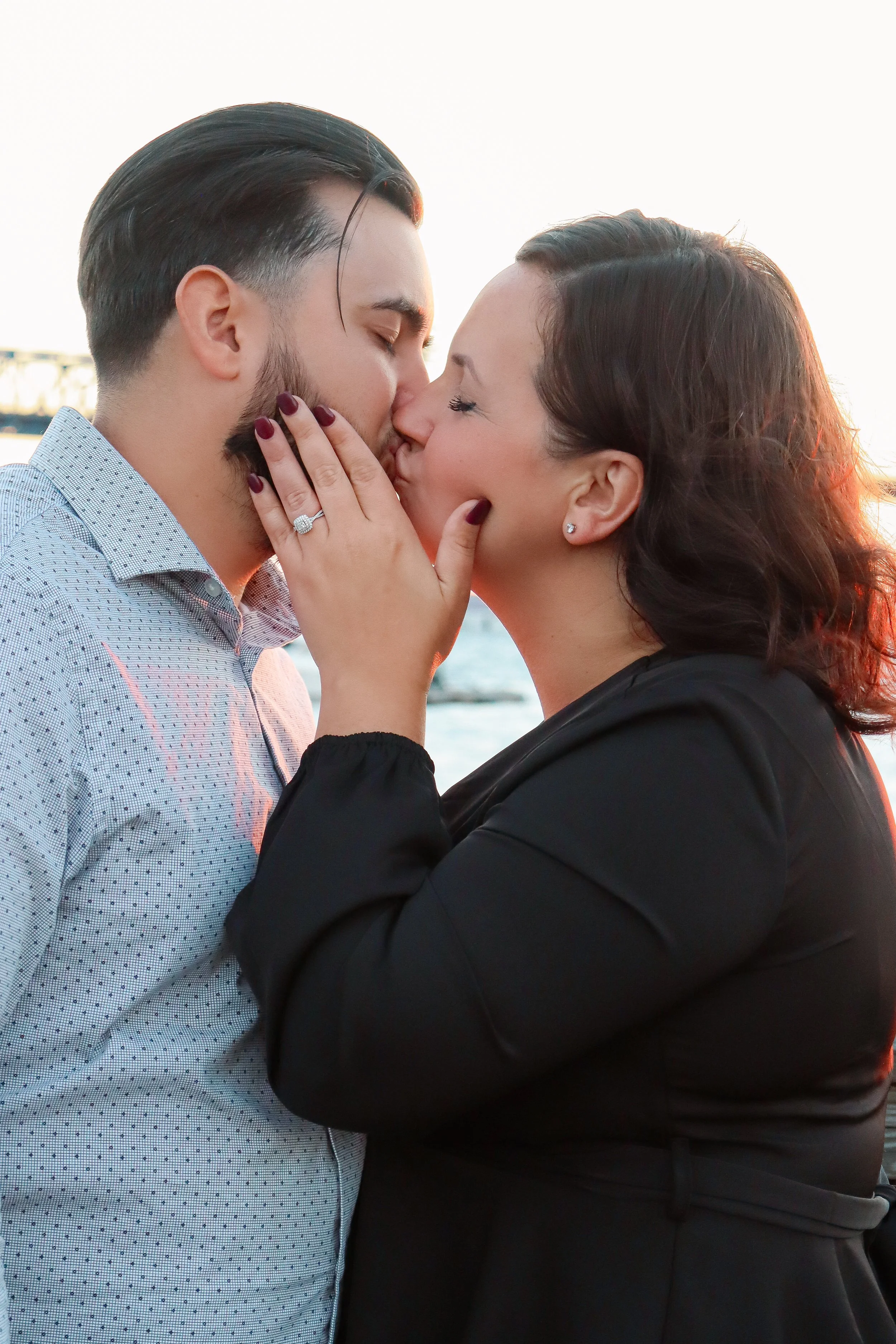 A couple sharing a kiss outdoors during sunset, with water and a bridge in the background.