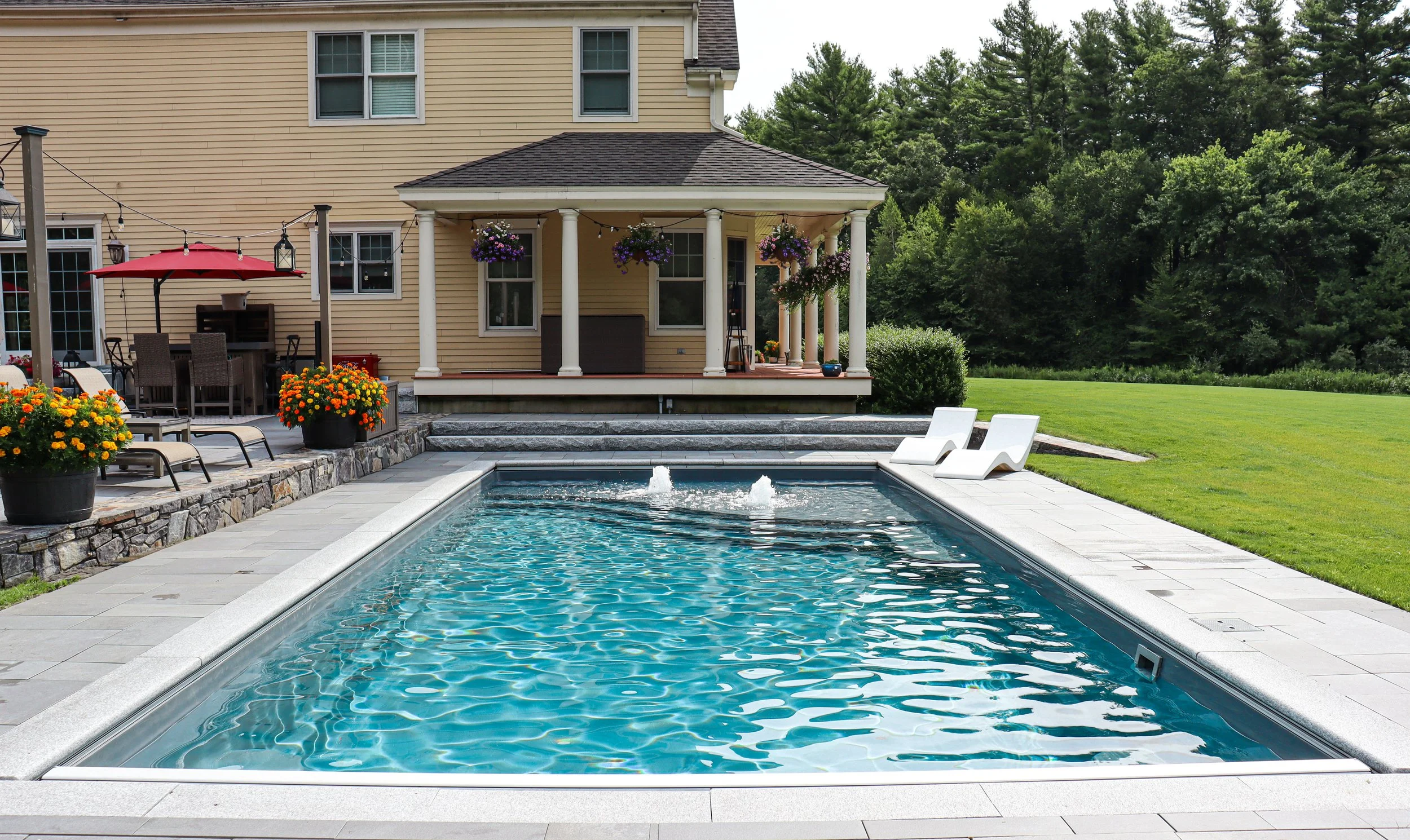 Backyard with swimming pool, lounge chairs, potted flowers, outdoor dining, and house with front porch, surrounded by green trees.