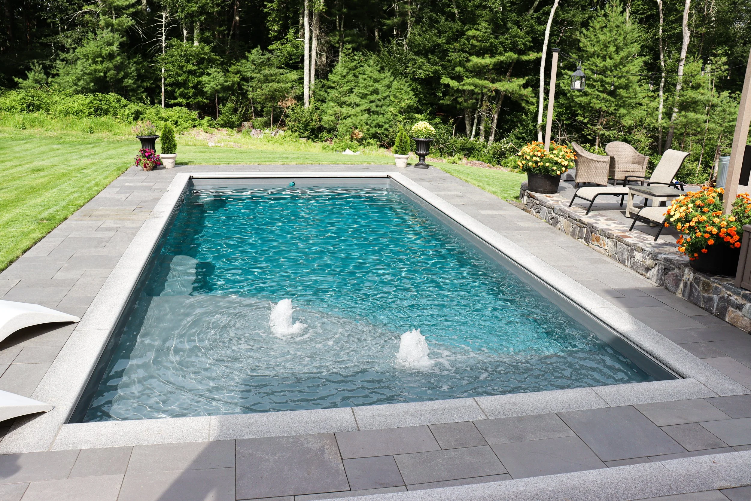Backyard swimming pool surrounded by a stone patio, with outdoor furniture and potted flowers, set against a background of green trees.