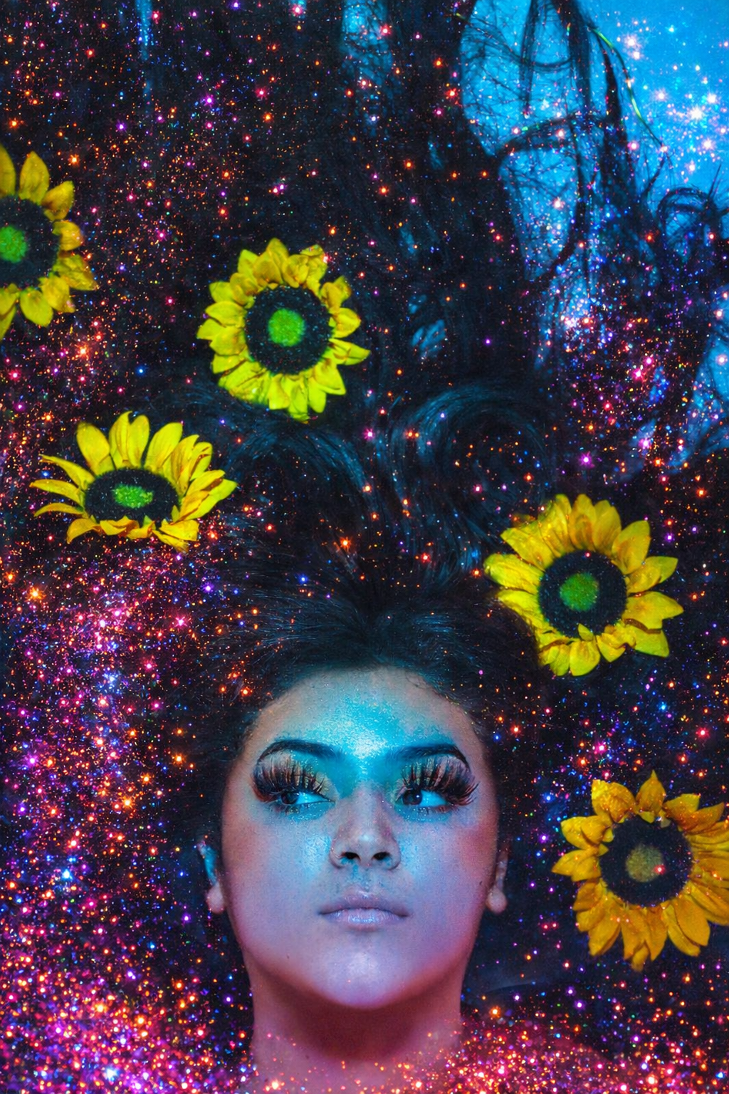 A woman with long, dark hair and makeup, lying down amidst colorful, glittering star-like particles and yellow sunflowers.