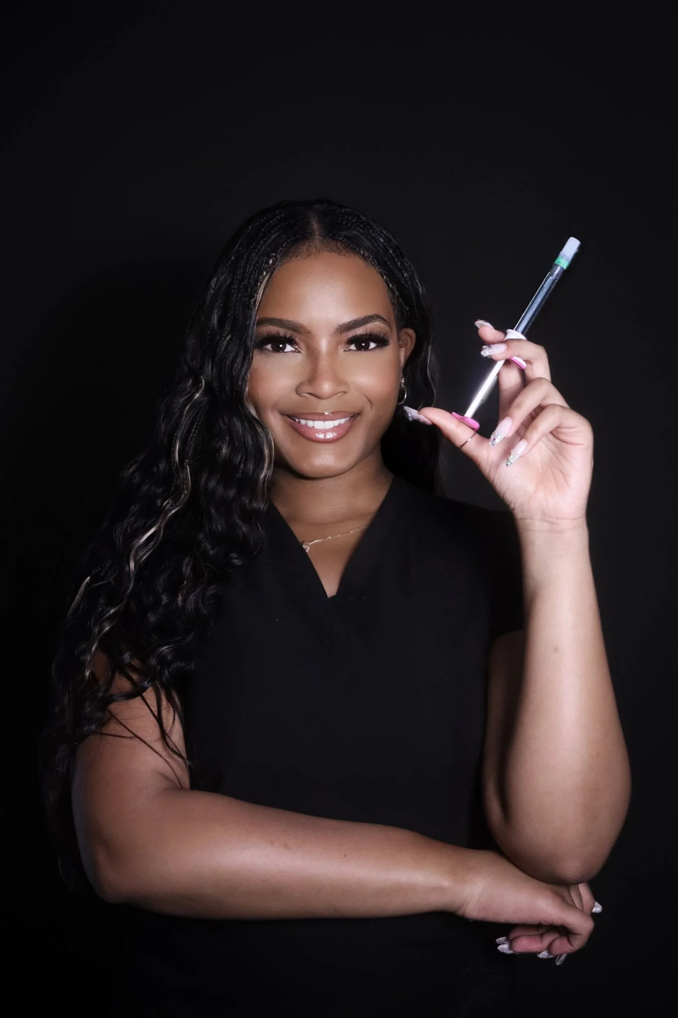 A woman with long, wavy black hair, smiling and holding a pipette, dressed in black against a dark background.